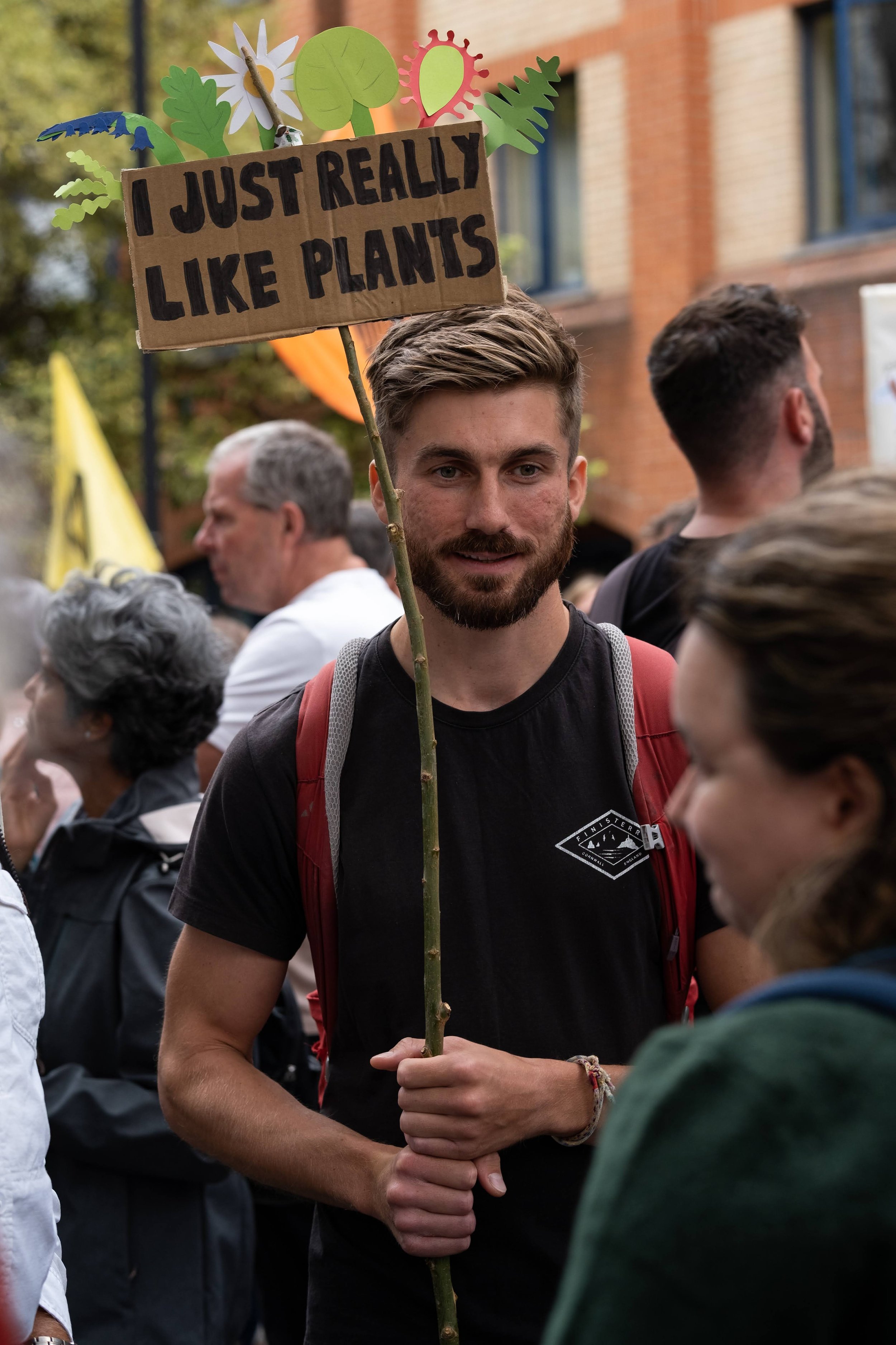 A young man with a beard holding a sign that reads "I just really like plants" at an outdoor protest or rally, surrounded by other people.