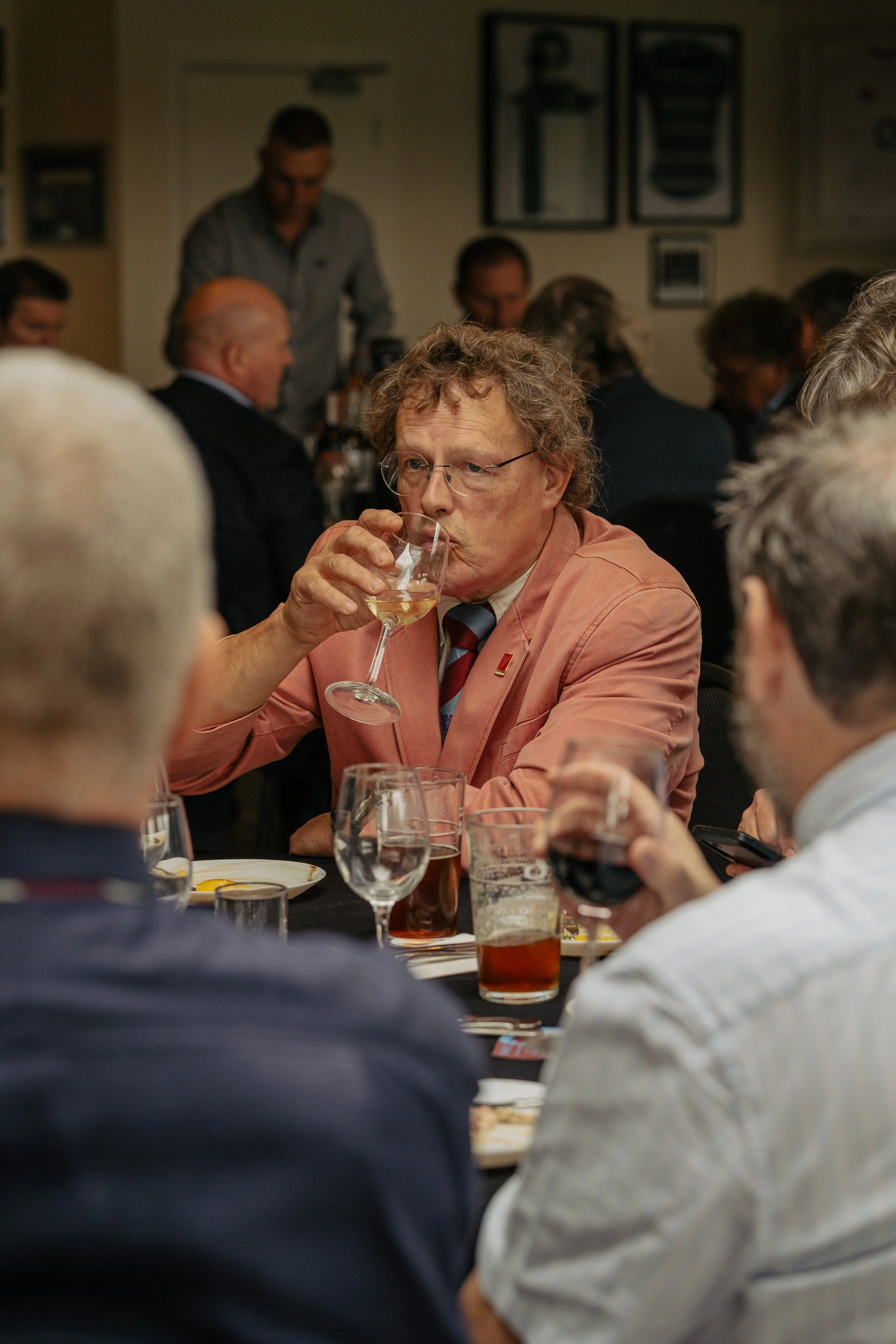 Man with glasses and curly hair in a pink jacket drinks white wine at a dinner gathering, surrounded by others with glasses of wine and beer, in a room with framed pictures.