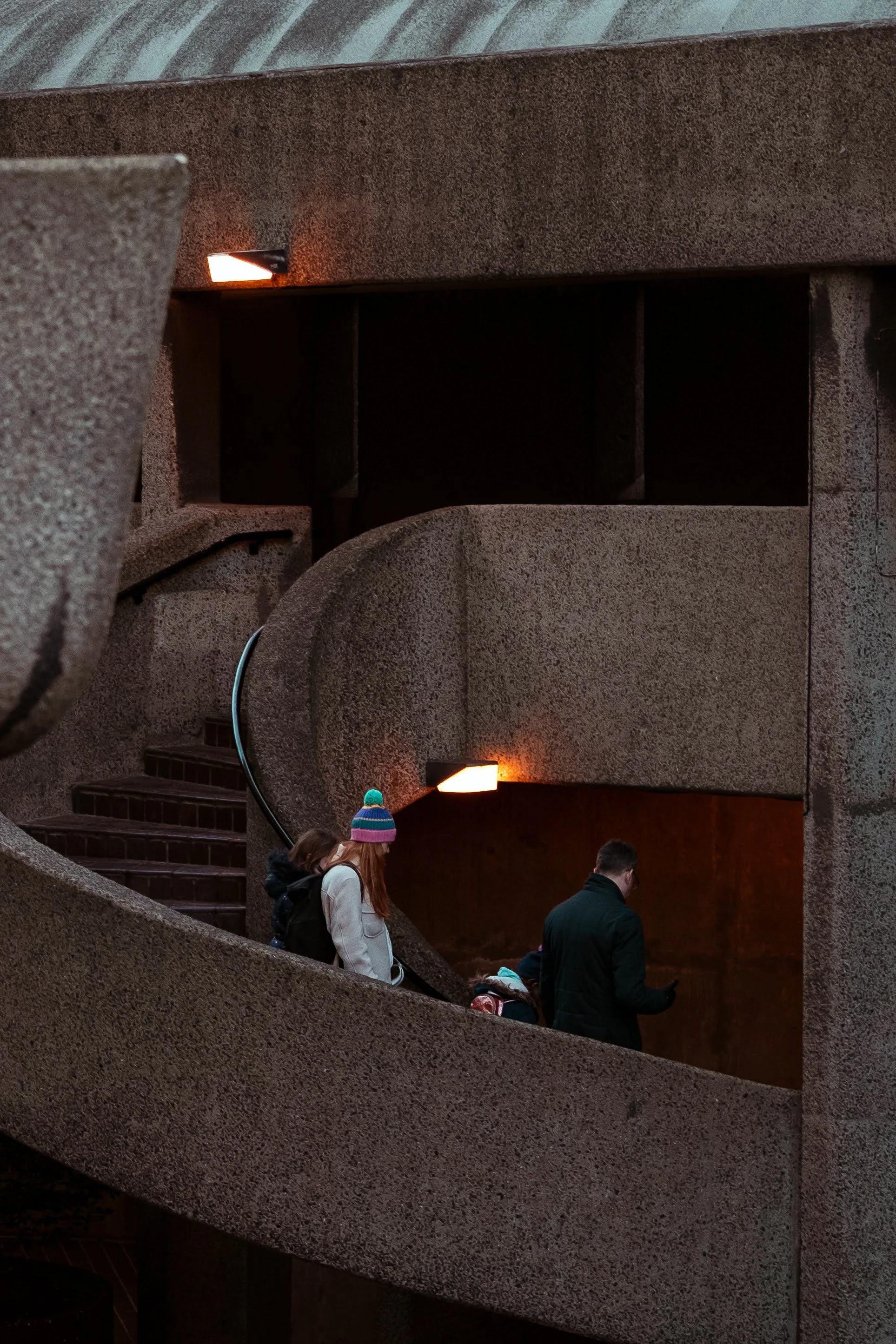People walking down an inclined concrete staircase inside a modern, textured building with warm lighting.
