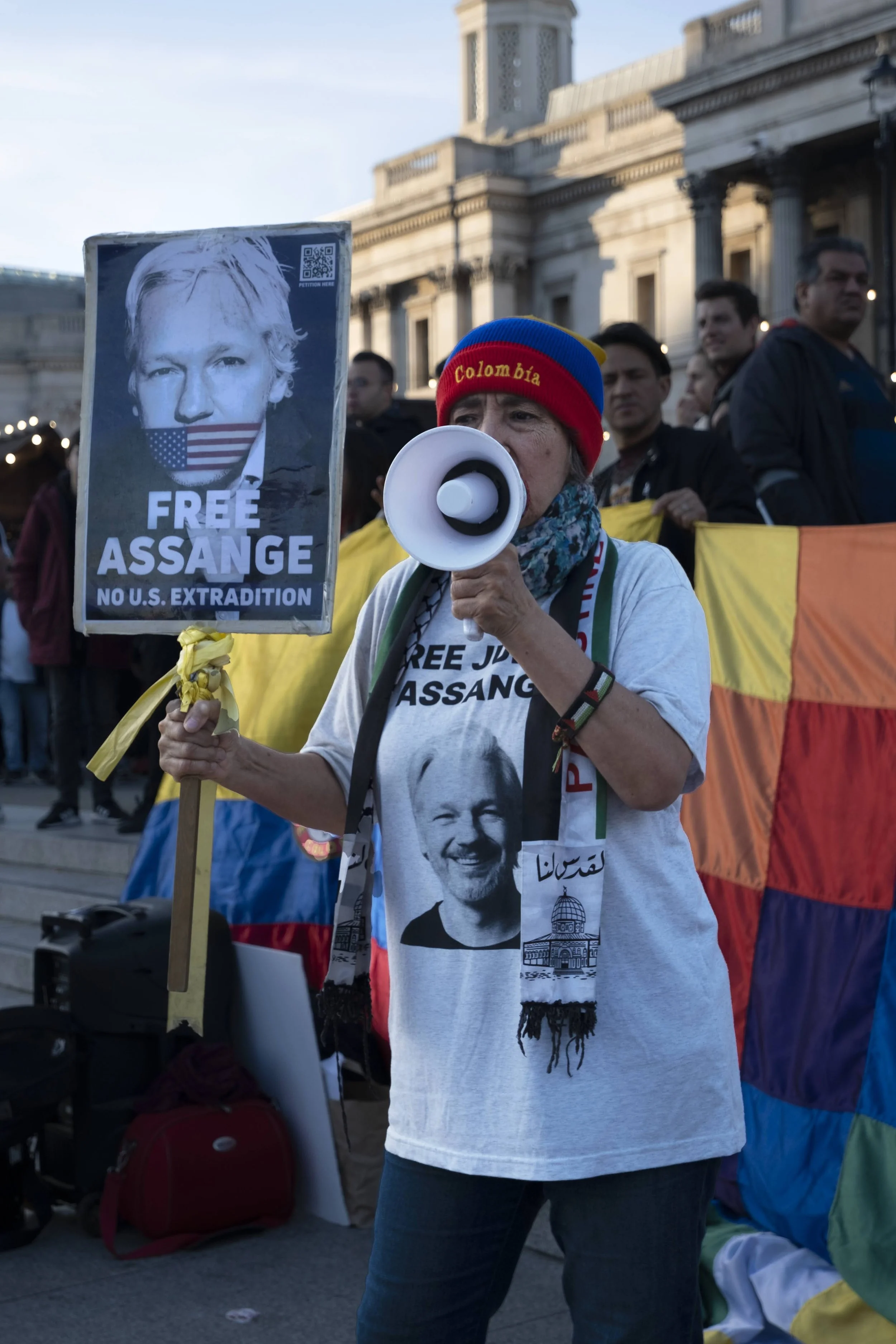 A woman at a protest holding a sign that says 'FREE ASSANGE NO U.S. EXTRADITION' with a picture of Julian Assange. She is wearing a beanie with the word 'Colombia' and a t-shirt with Assange's image. She is speaking into a megaphone.