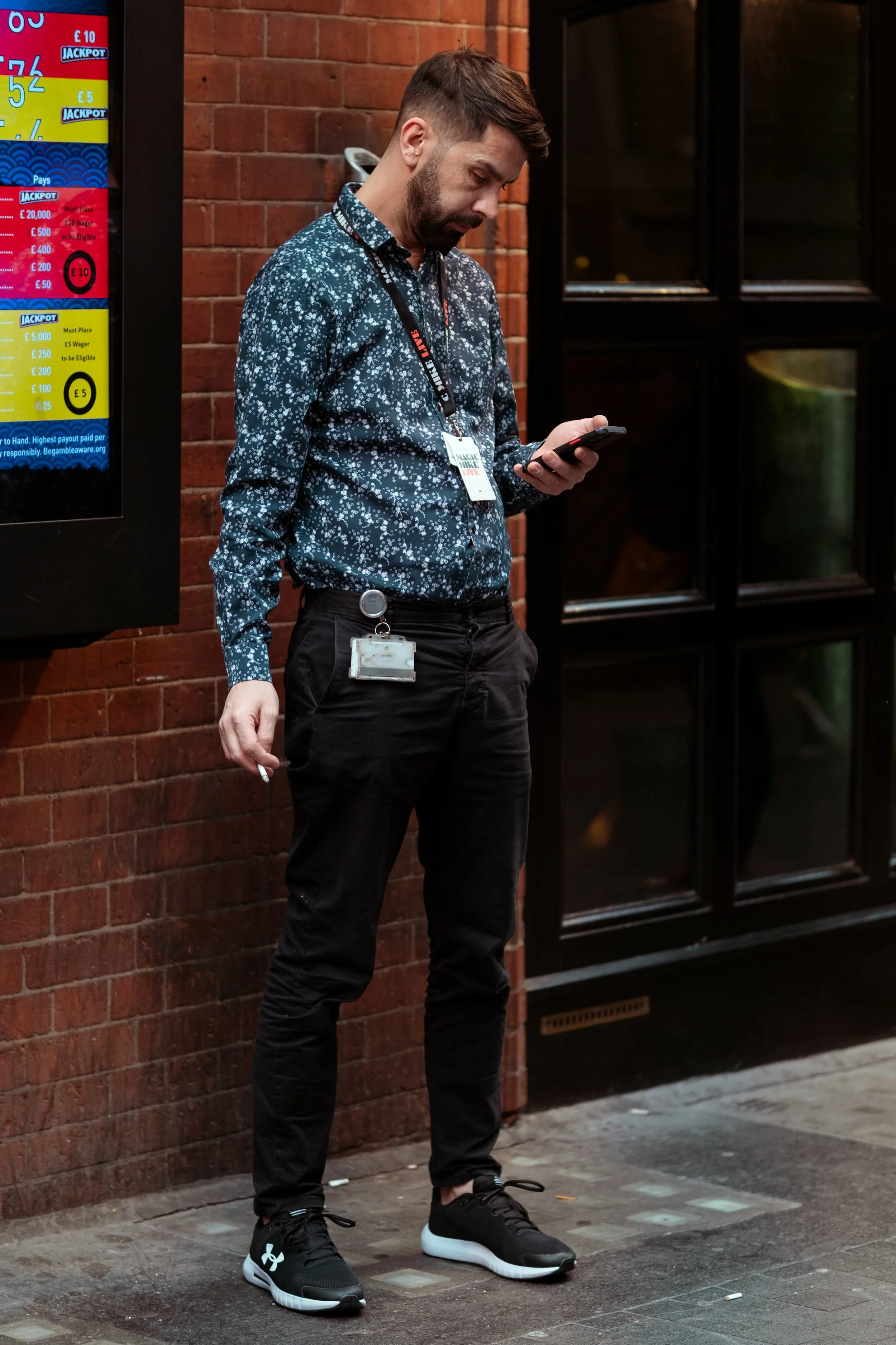 A man with a beard and mustache wearing a patterned blue shirt, black pants, and black athletic shoes stands on a sidewalk, looking at his smartphone near a red brick wall and a lottery jackpot sign.