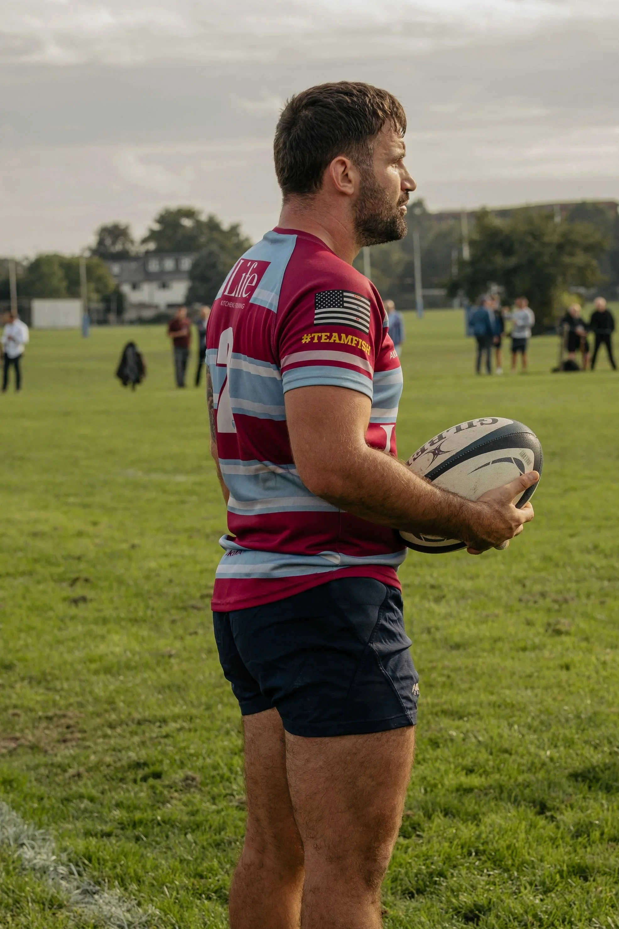 A rugby player in a maroon and light blue striped jersey holding a rugby ball on a grassy field with people in the background.