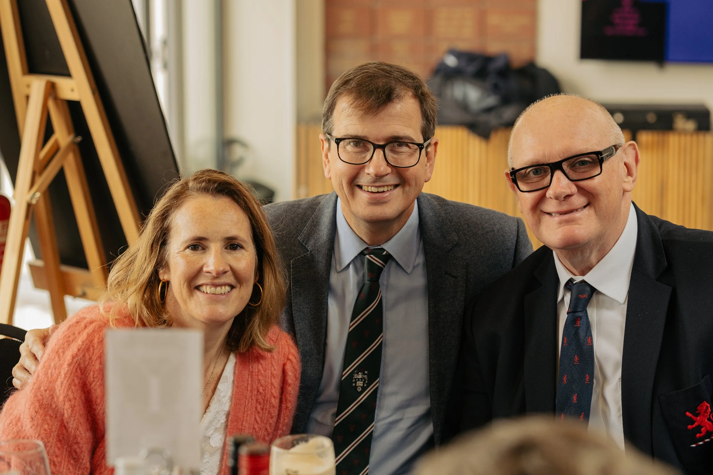 Three people smiling and posing for a photo at a social gathering or event, sitting at a table with drinks.