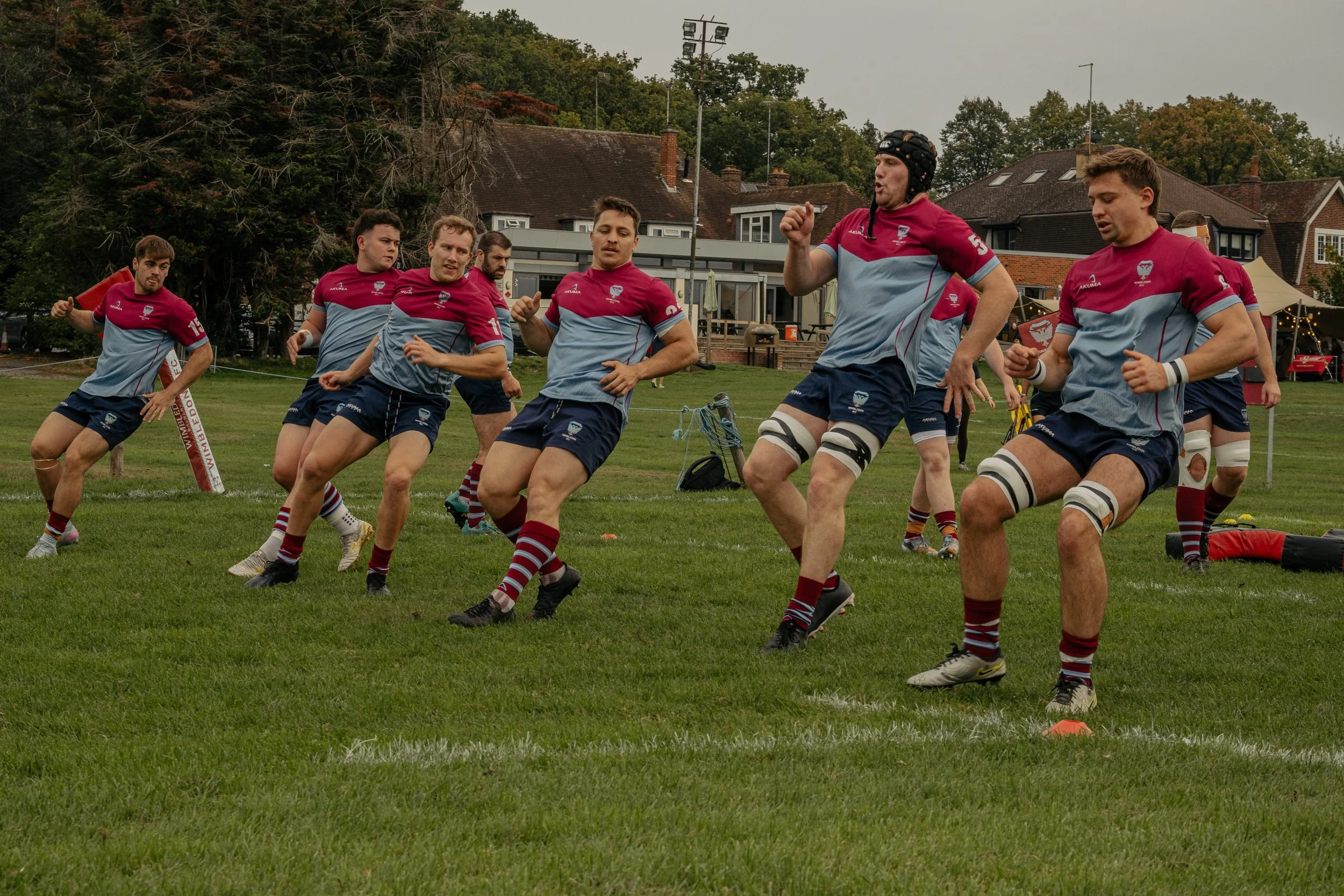 A rugby team practicing on a grassy field, wearing matching maroon and grey uniforms, with some players wearing protective gear, and a few of them engaged in a drill or warm-up exercise.