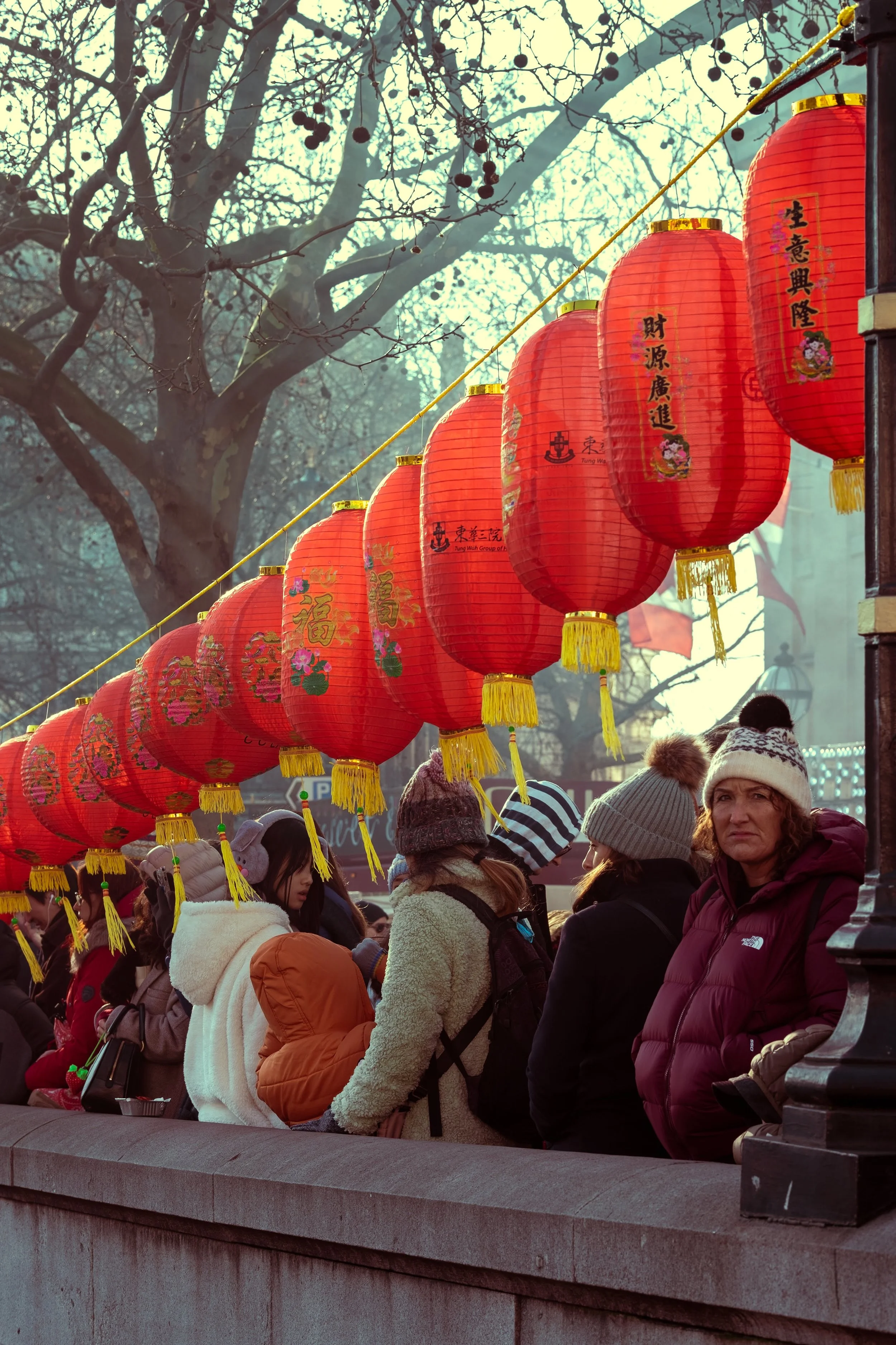People wearing winter clothing gather behind a row of red Chinese lanterns strung along a street, with bare trees in the background.