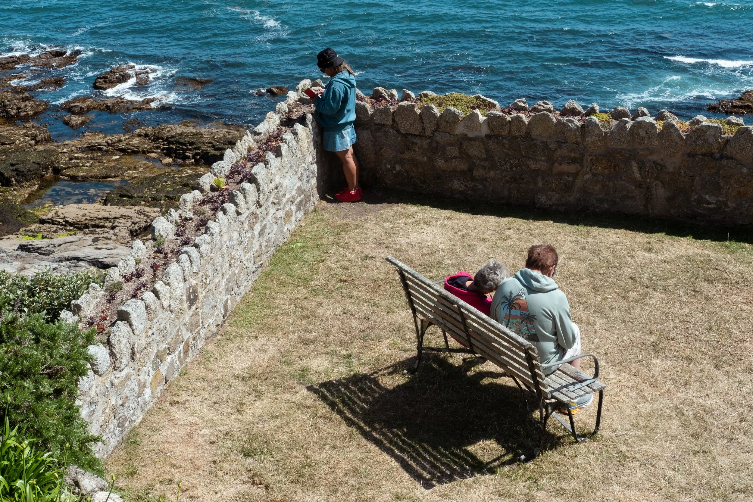 A woman and a young girl are by a stone wall overlooking the ocean. A man and an older woman are sitting on a wooden bench nearby on a grassy area.