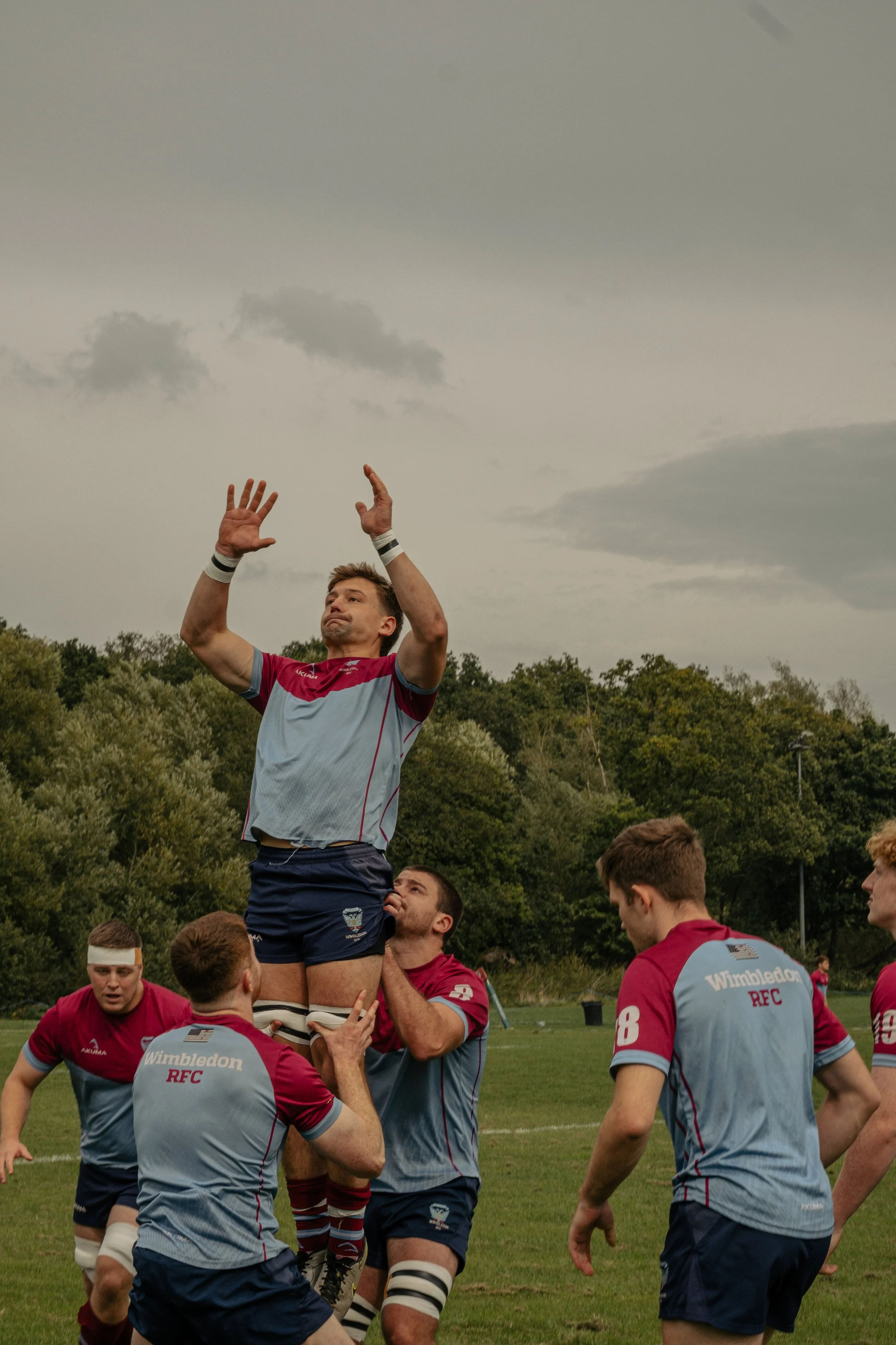 A rugby player being lifted by teammates during a line-out, wearing a jersey with Manchester United's logo, on a field with trees and a cloudy sky in the background.