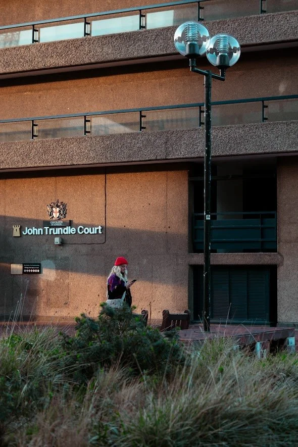 A woman wearing a red hat and purple jacket is standing outdoors near a building, looking at her phone, with a bench and some grass in the foreground. The building has a sign that reads "John Trundle Court" and features a crest or emblem above the si