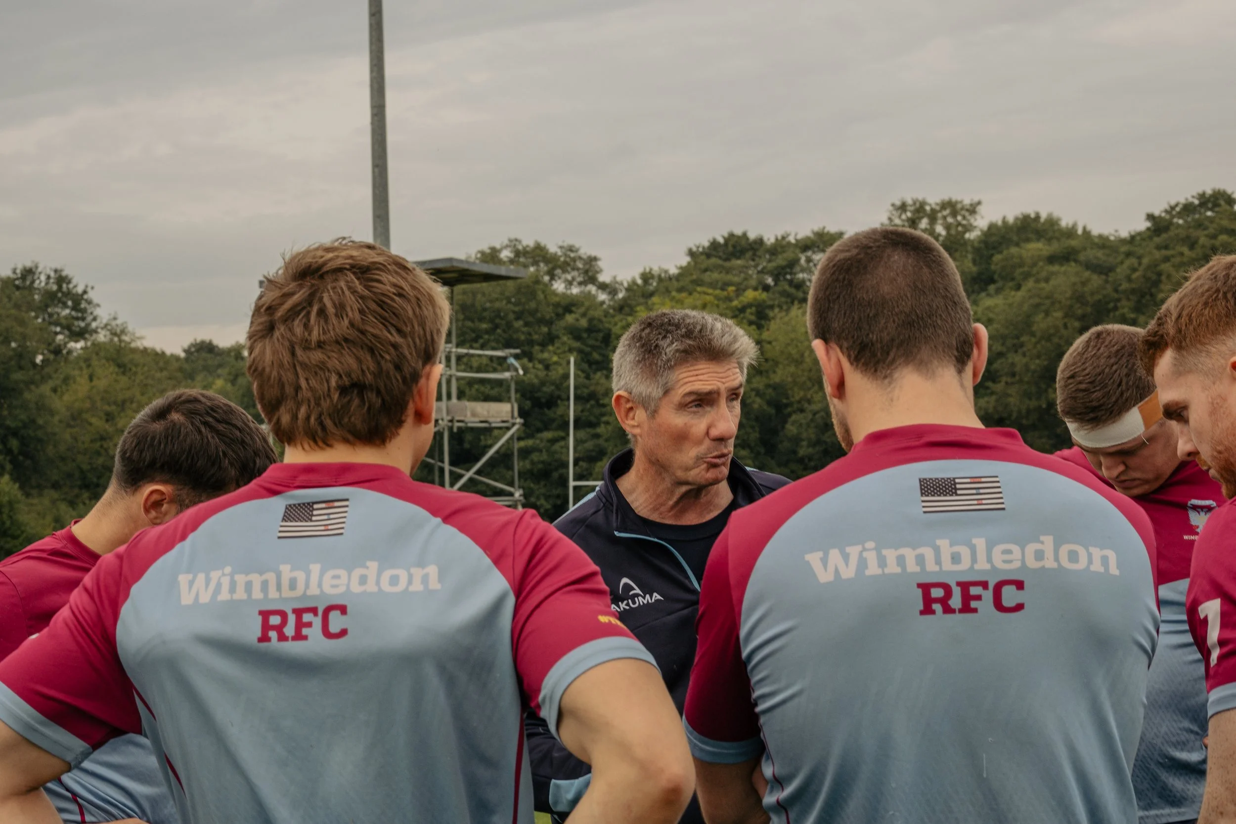 Rugby coach giving instructions to a team of players during a practice at an outdoor field, with trees and a cloudy sky in the background.