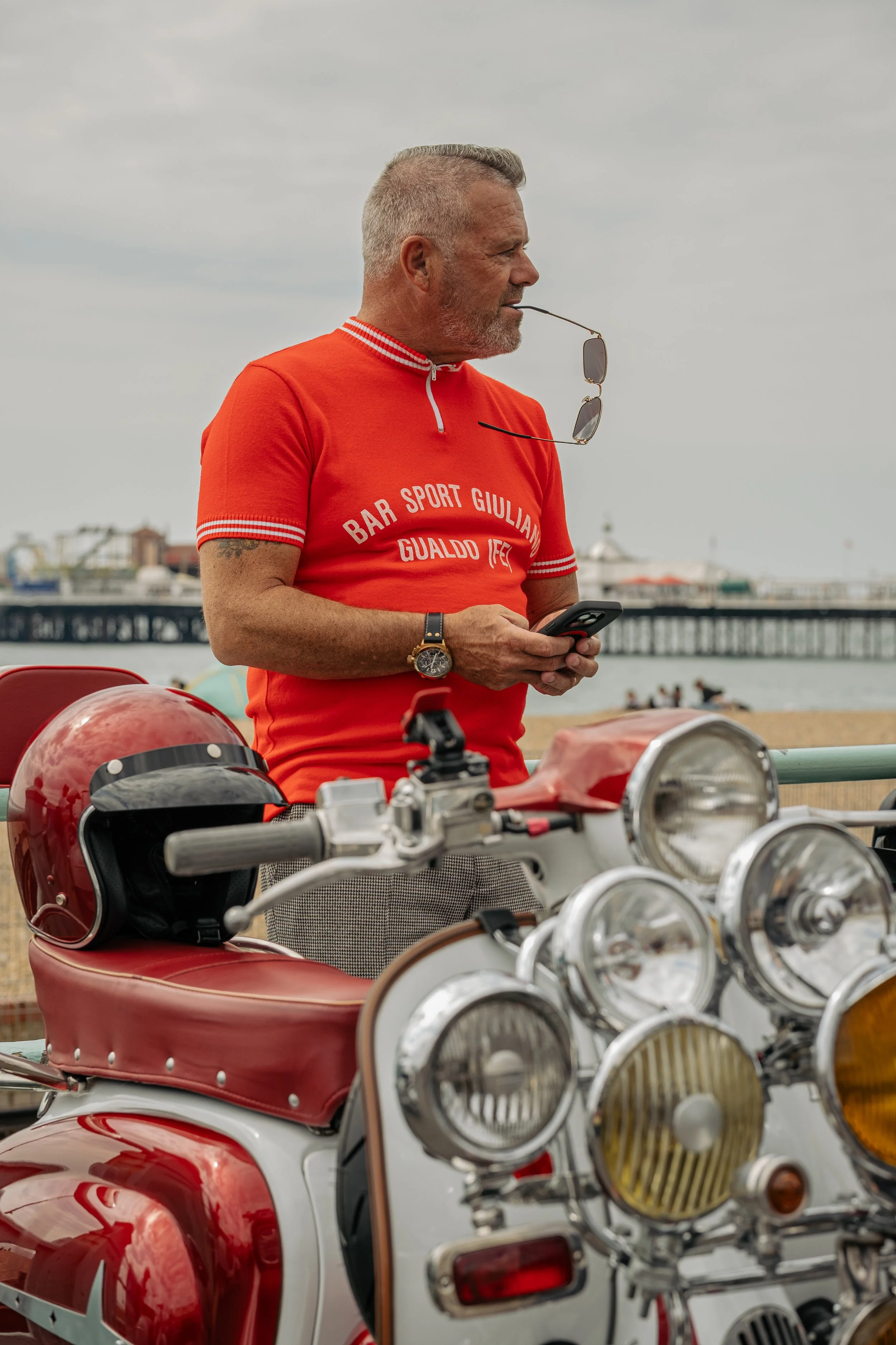 A middle-aged man with gray hair and a beard stands near a white and red vintage scooter, wearing an orange T-shirt with white text, holding a smartphone, with sunglasses hanging from his shirt collar, a black watch on his left wrist, and a red helme