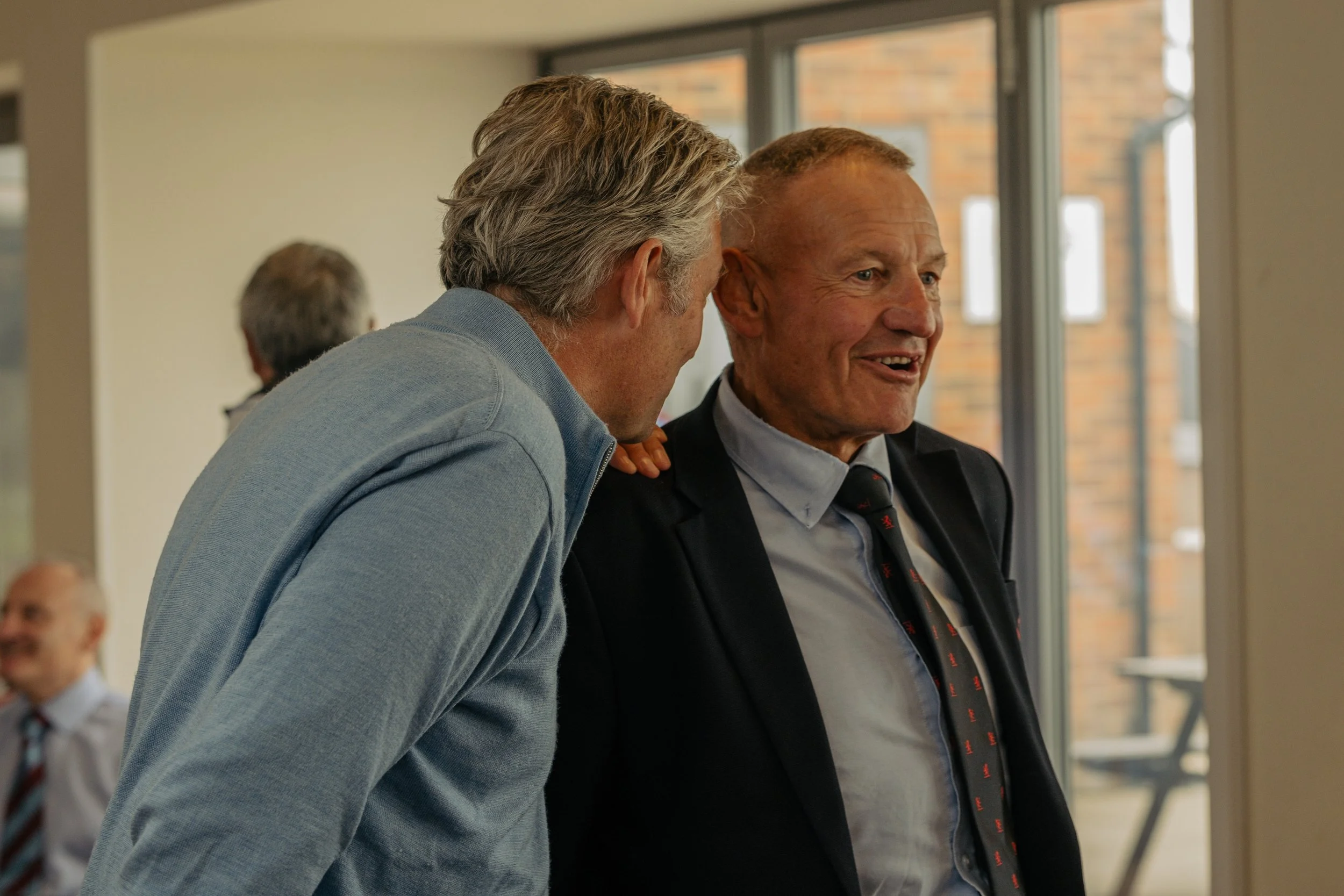 Two middle-aged men are engaged in conversation during a social gathering indoors, with large windows in the background. One man has gray hair and is wearing a light blue zip-up sweater, while the other is bald and wearing a black blazer and tie.
