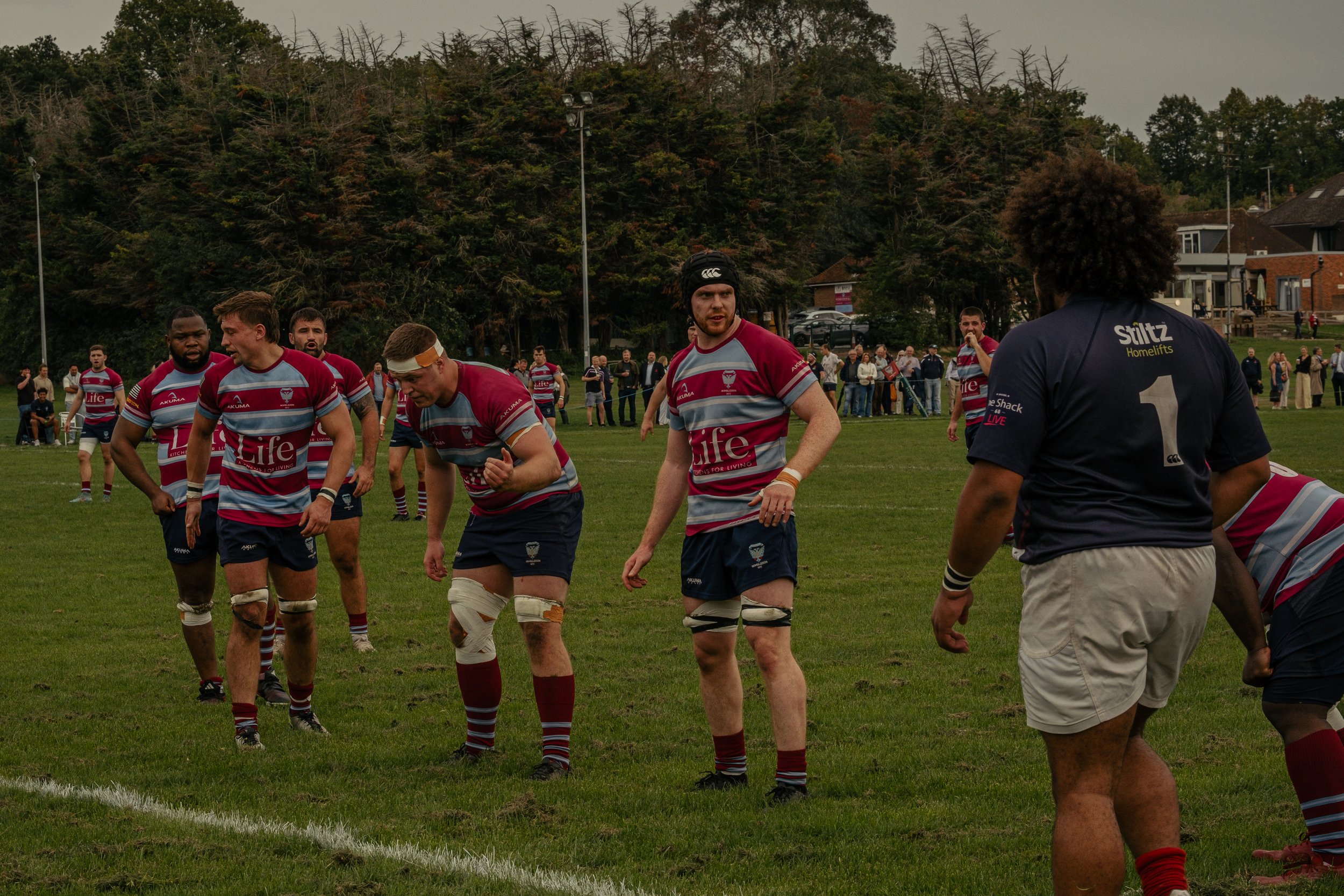 Rugby players on the field during a match with spectators watching from the sidelines and houses in the background.