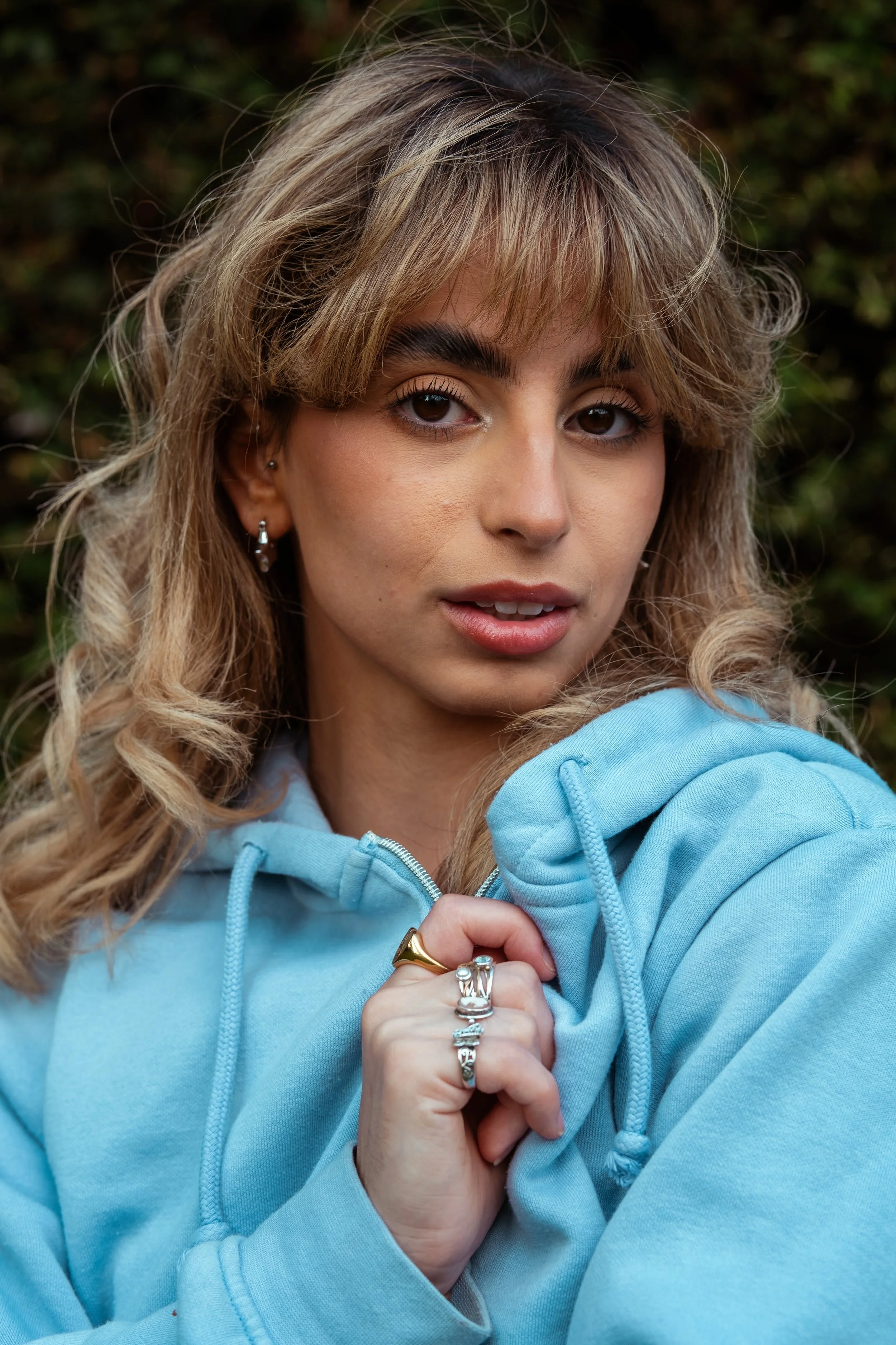 A young woman with curly blonde hair and earrings wearing a light blue hoodie, looking at the camera with a neutral expression, outdoors with a blurred green background.