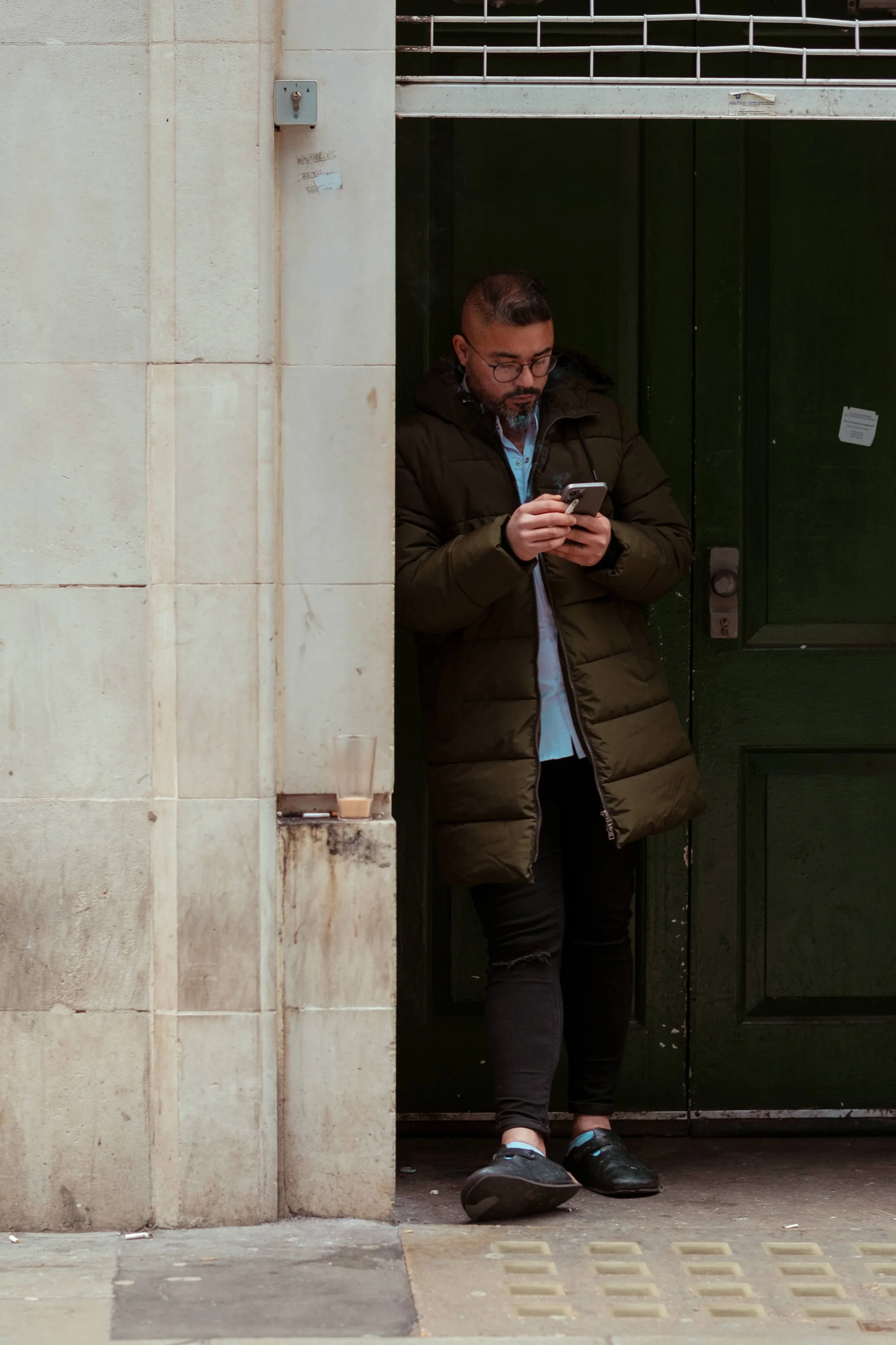 A man standing at a doorway, wearing a dark green jacket and glasses, looking at his phone. There is a half-empty glass of beverage placed on a ledge near him.