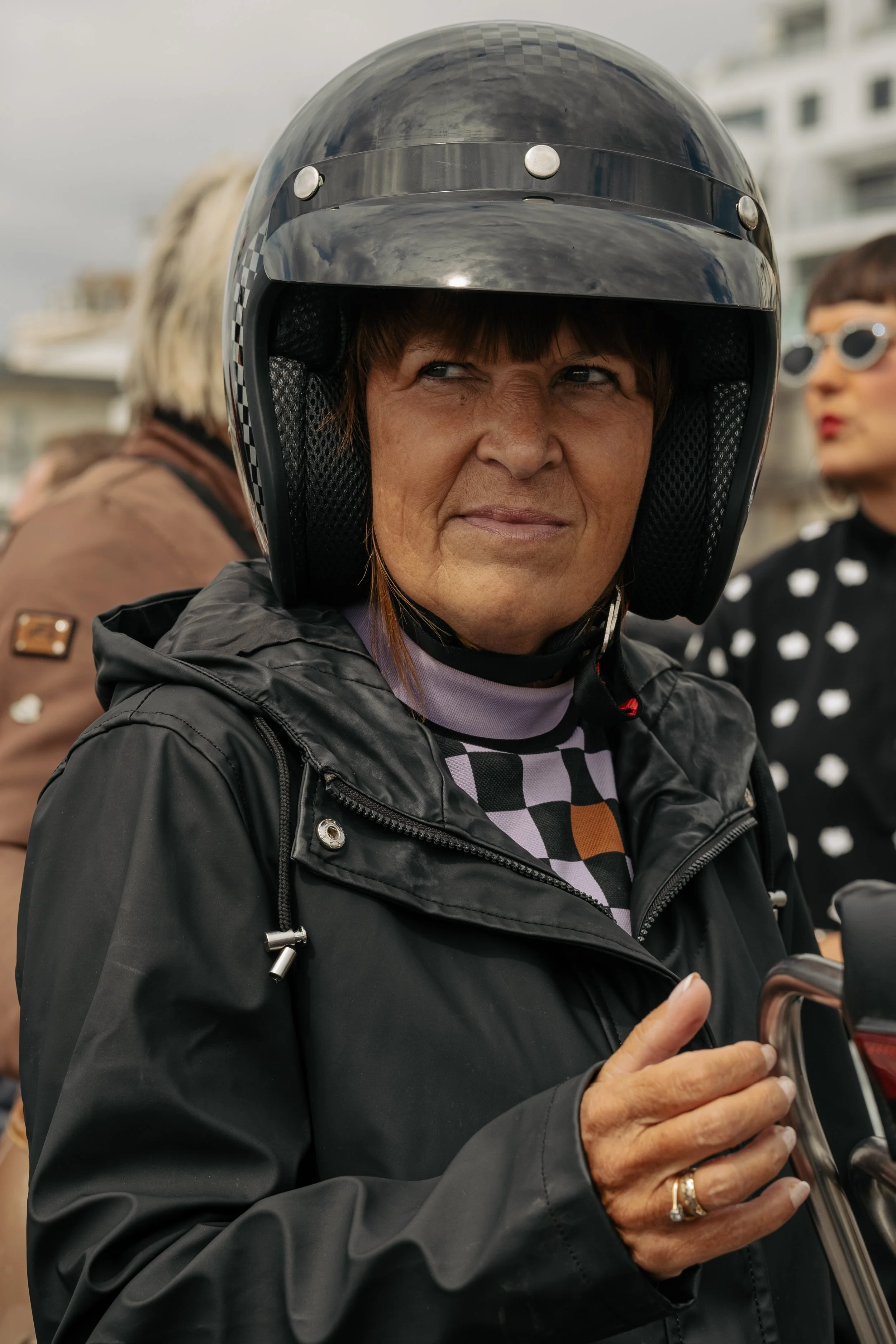A woman wearing a black motorcycle helmet and black jacket standing outdoors with other people in the background.