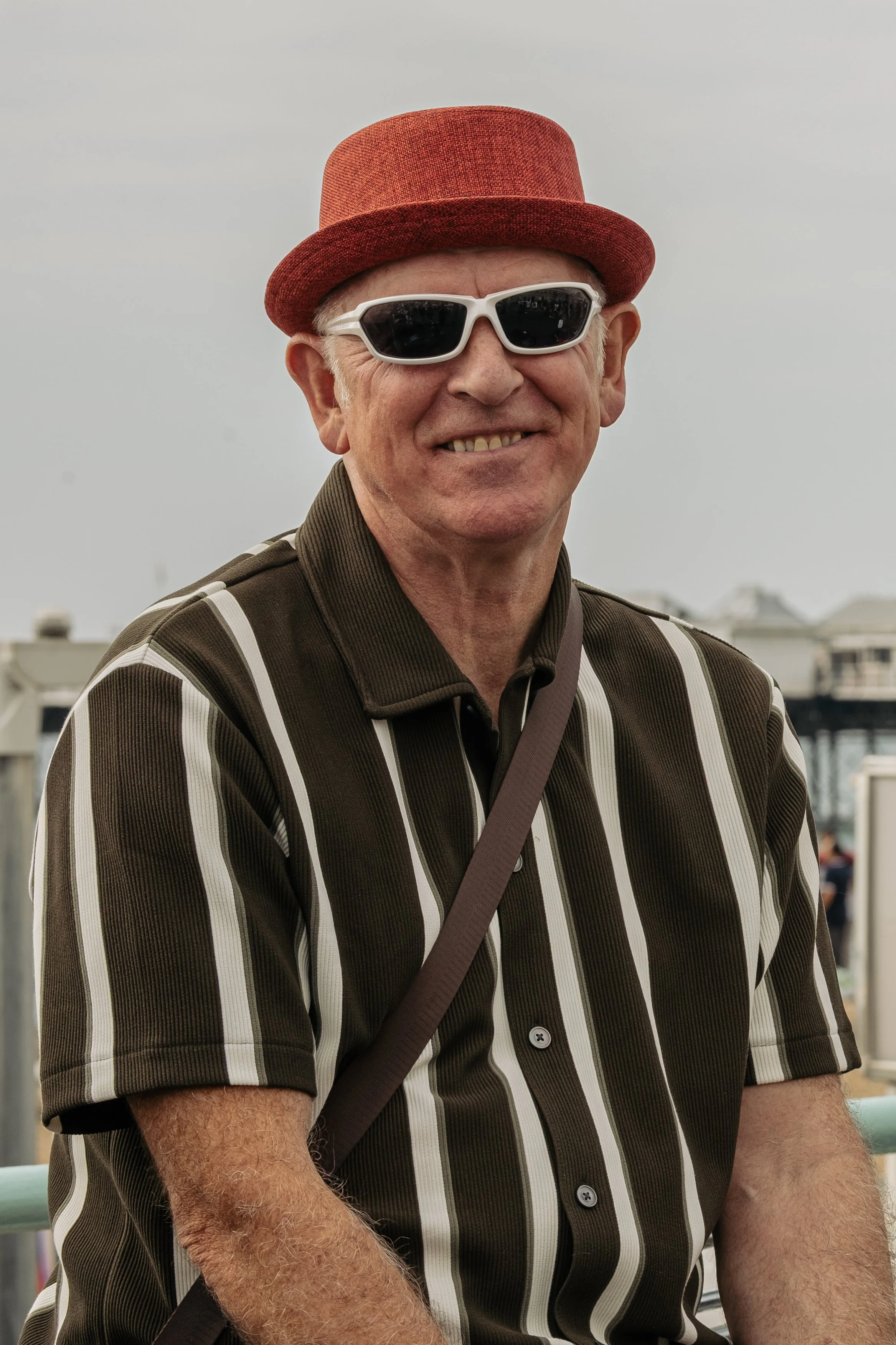 A smiling elderly man wearing a red hat, white sunglasses, a striped brown and white short-sleeve shirt, and a brown strap over his shoulder, outdoors with cloudy sky in the background.