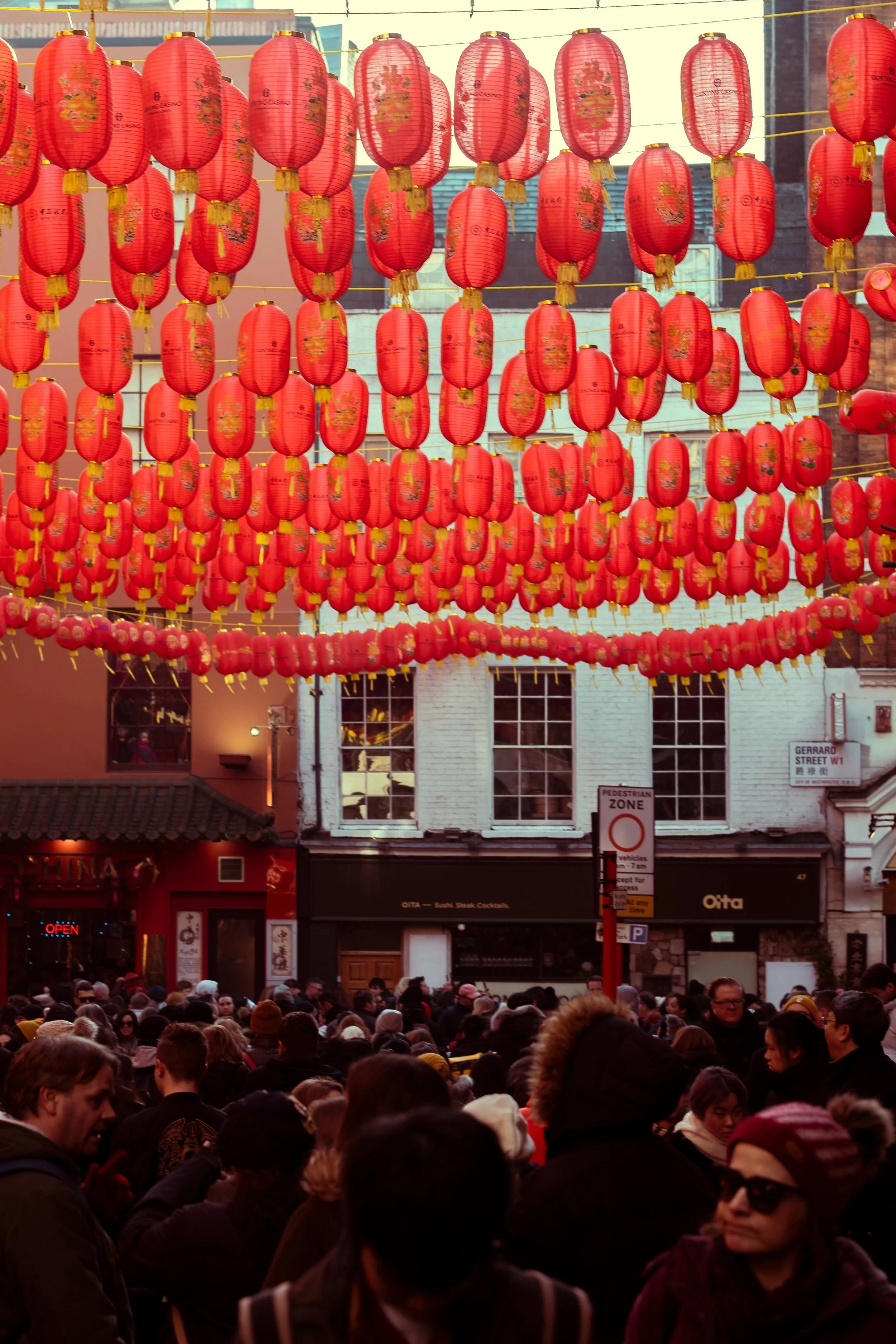 A street scene decorated with multiple rows of red Chinese lanterns hanging above a crowd of people, with storefronts in the background.
