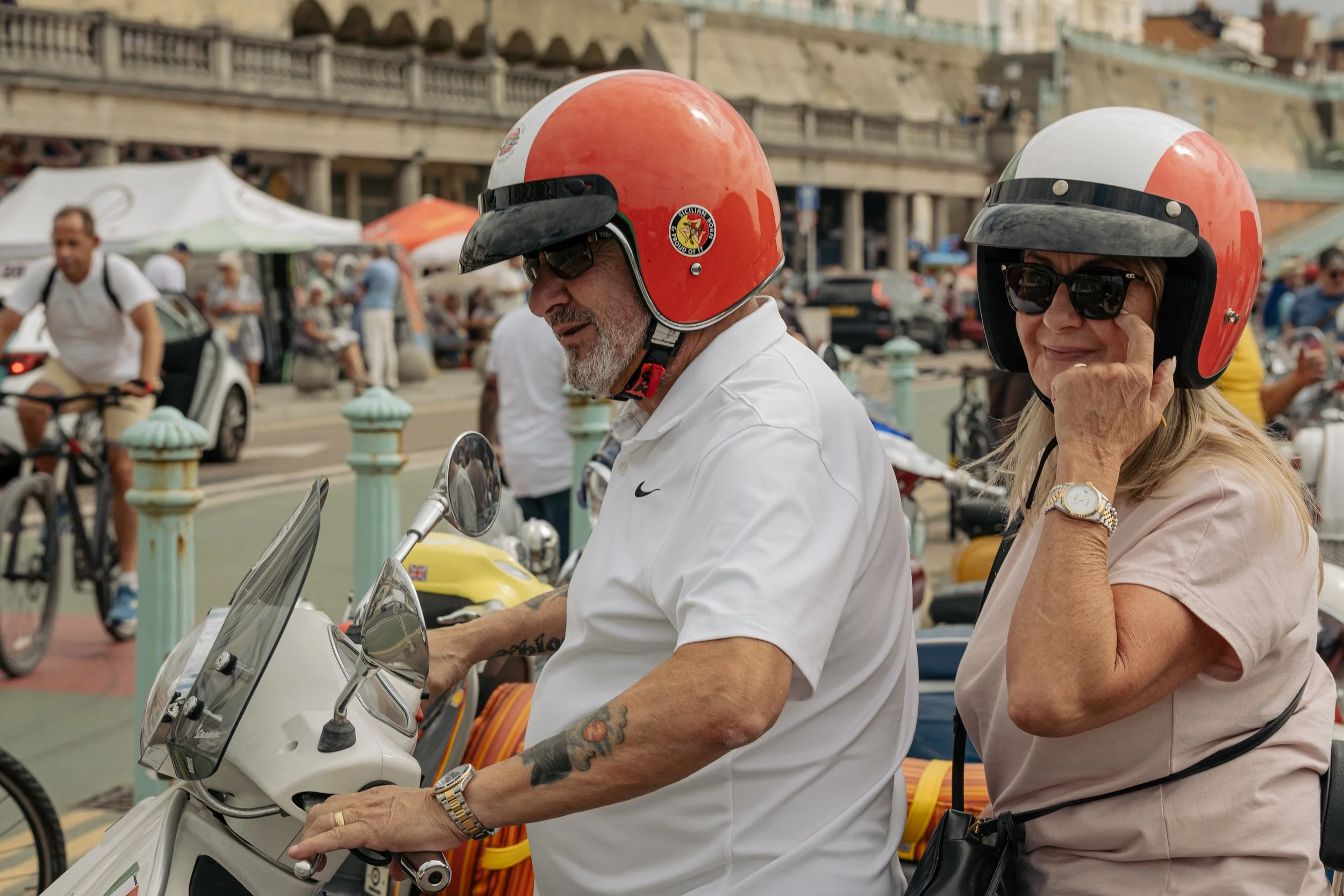 Two older adults, a man and a woman, wearing red helmets and sunglasses, riding a scooter at a lively outdoor market or event with tents, vendors, and other people in the background.