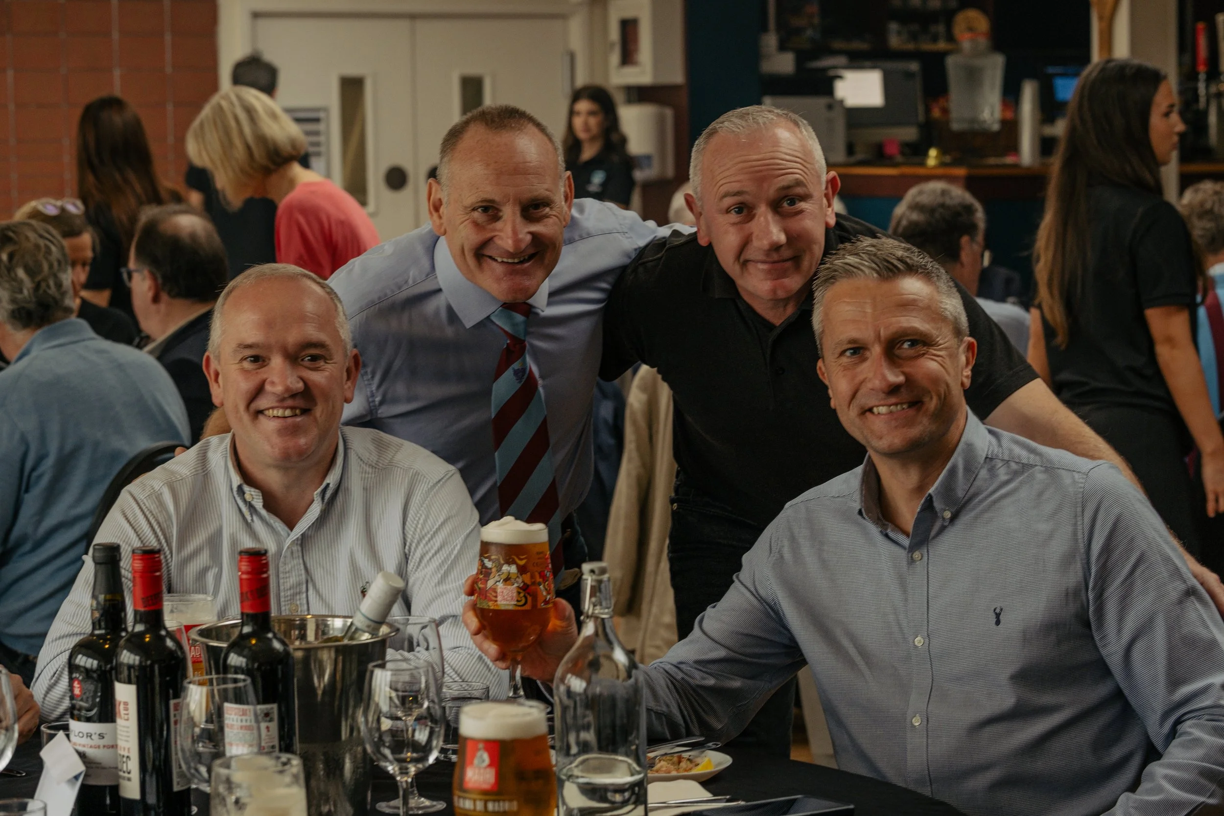 Four men gathered around a dinner table at a social event, smiling at the camera, with drinks and bottles on the table. In the background, other guests are mingling and talking.