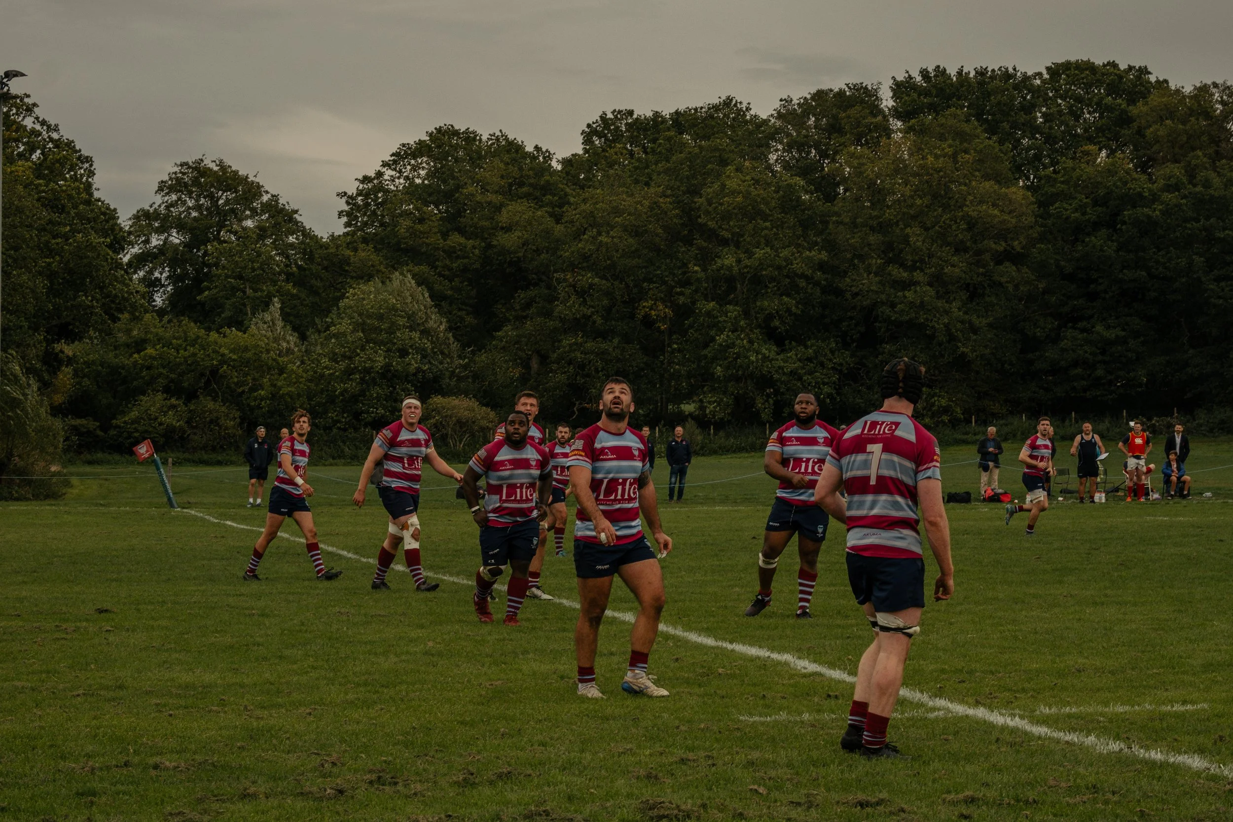 Rugby players on the field during a match, with some players running and others standing, under a cloudy sky, with spectators and staff in the background.