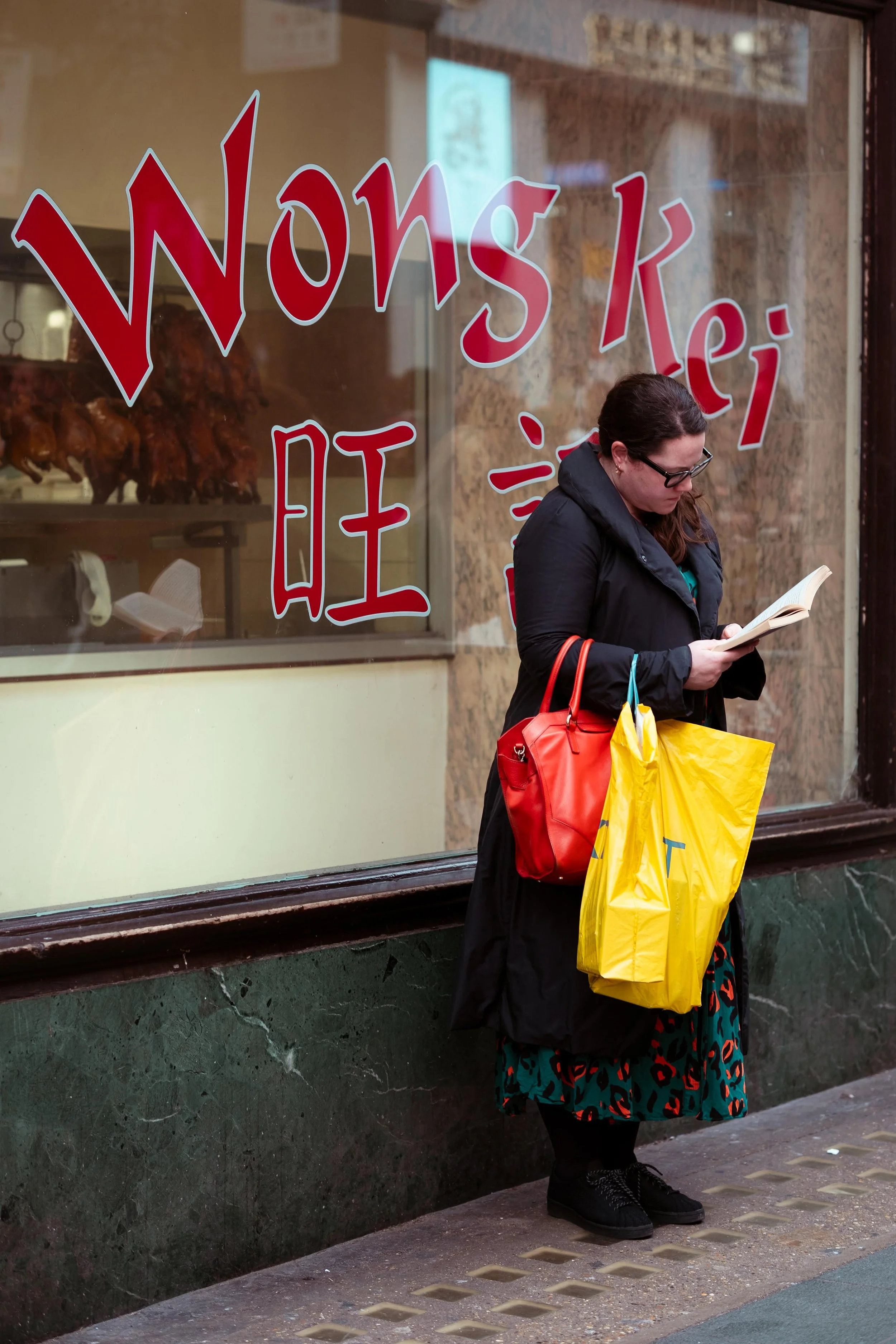 A woman standing on a city sidewalk reading a book outside a restaurant or shop with large red and white Chinese characters on the window. She is carrying two shopping bags and wearing glasses, a dark coat, and black shoes.
