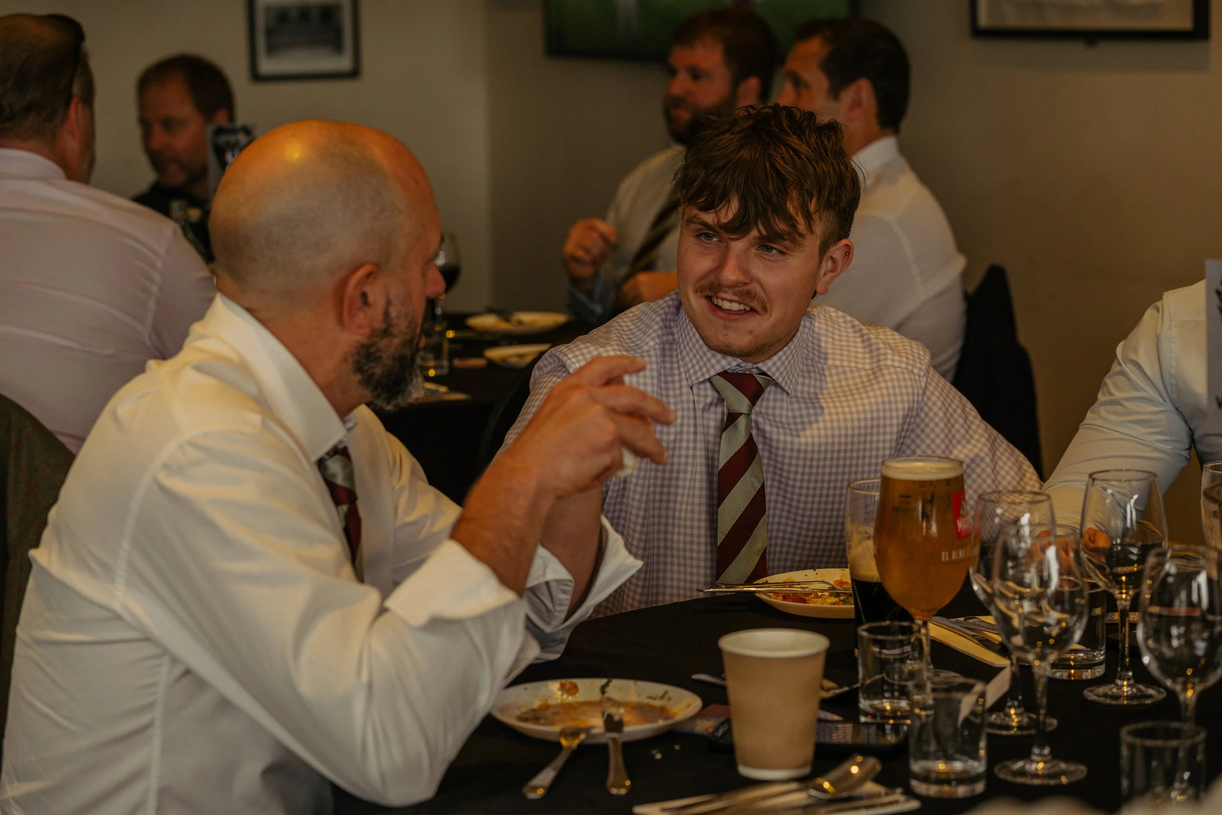 Two men in formal attire, sitting at a dinner table, engaged in conversation, with other people in the background.