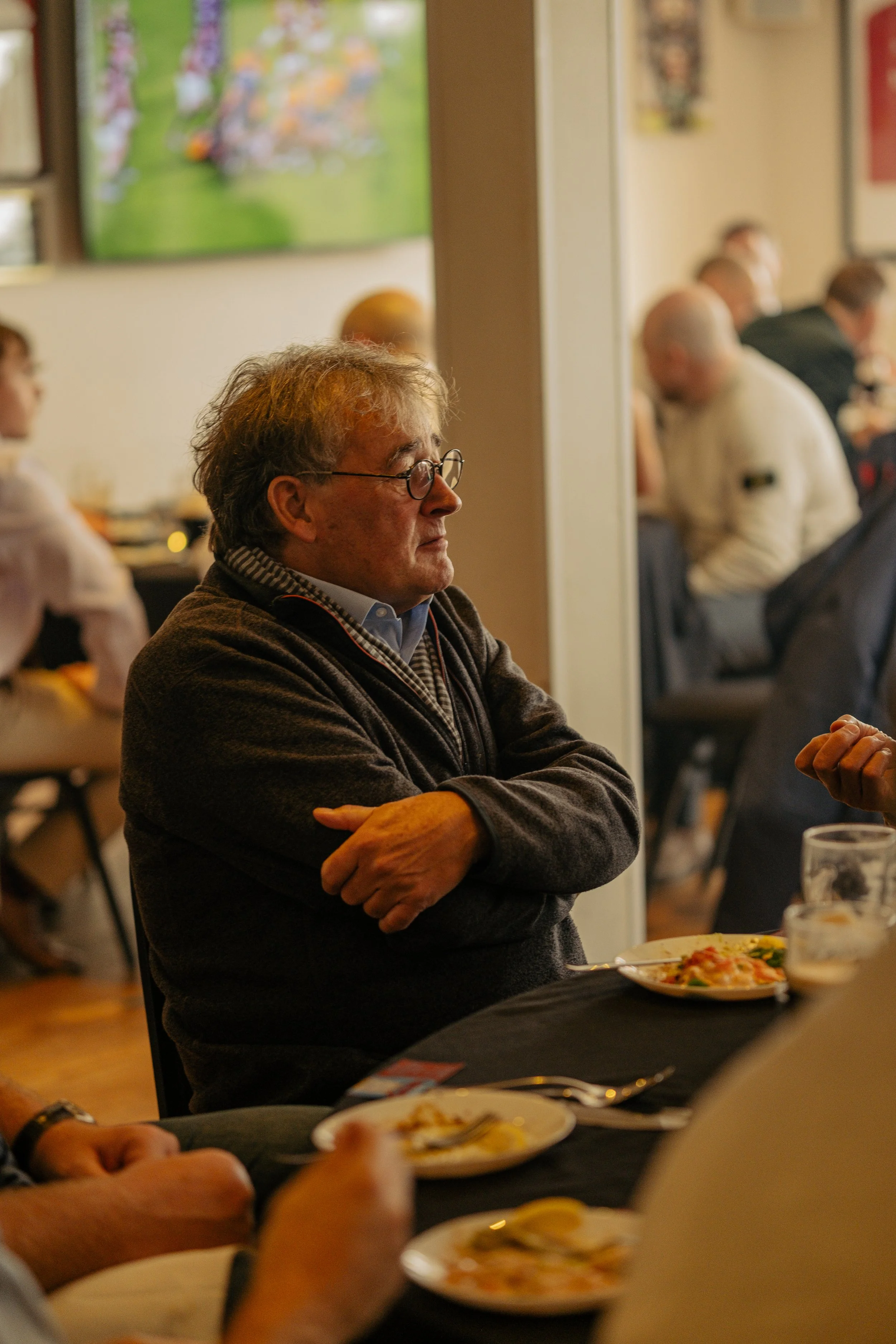 A middle-aged man with glasses and wavy hair sitting at a table with crossed arms, wearing a dark sweater, in a restaurant or dining area with other diners in the background.