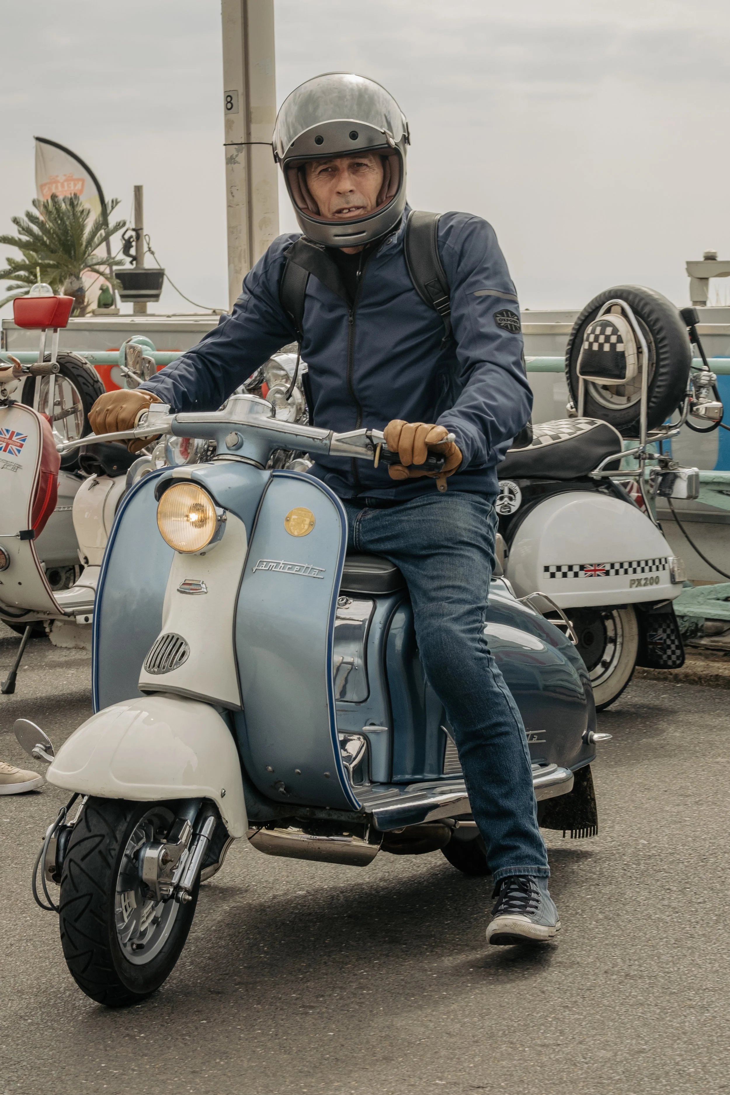 A man riding a vintage blue and white scooter, wearing a helmet, leather gloves, a navy jacket, and jeans, with other scooters in the background at an outdoor location.