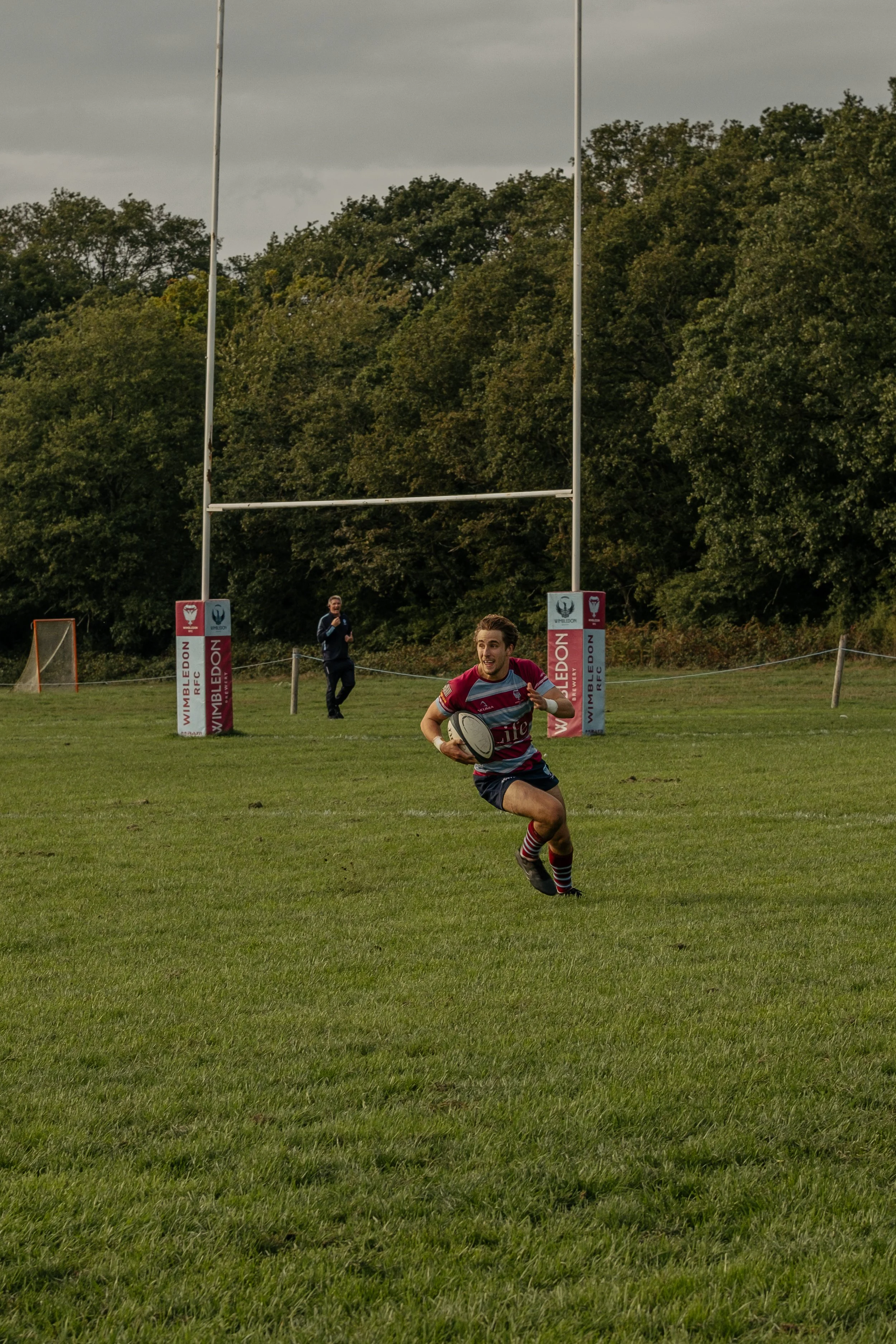A rugby player running on a grassy field while holding a rugby ball, with goal posts and some trees in the background.