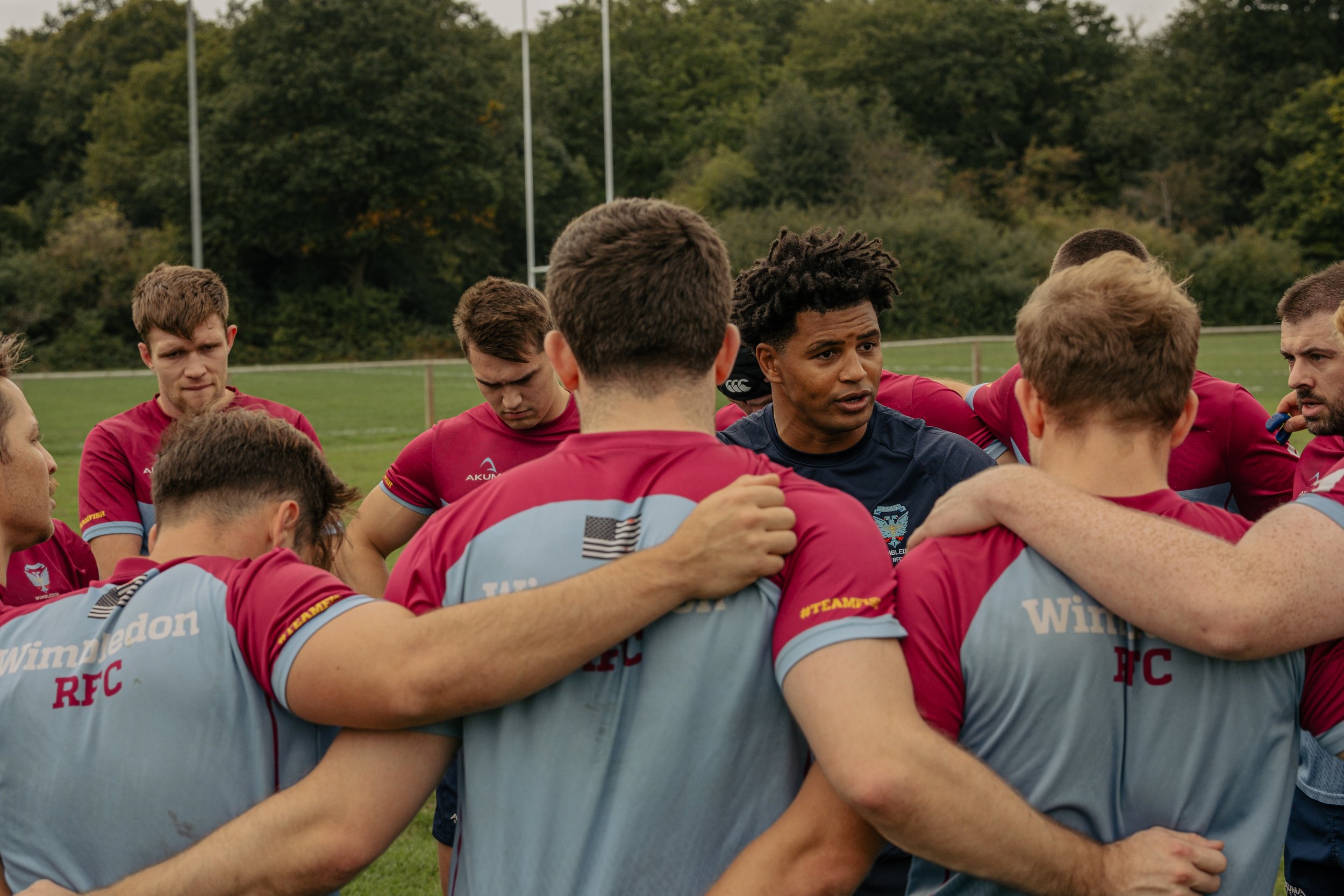 A rugby team huddles together on a field, listening to their coach during a game or practice, with trees in the background.