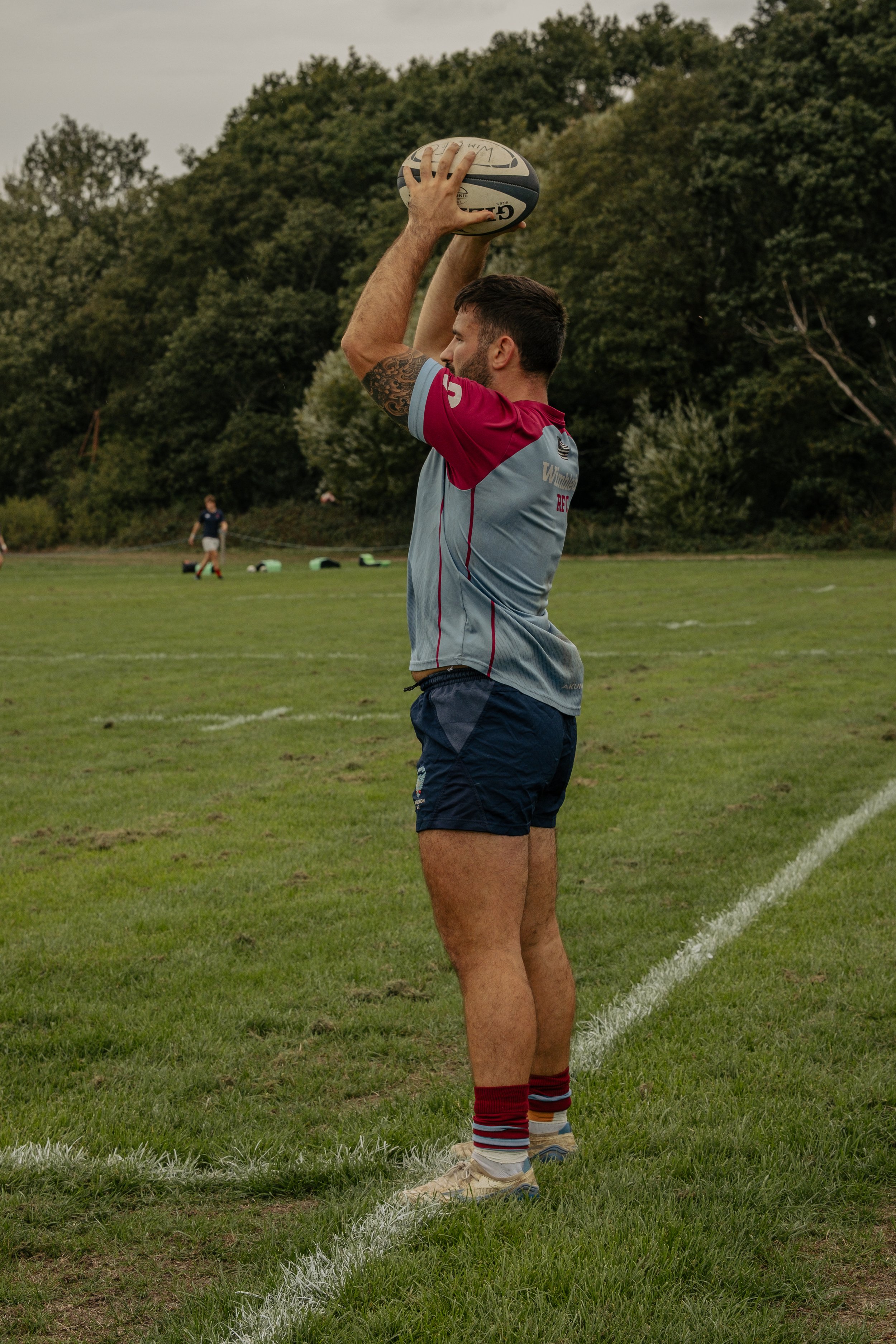 A rugby player holding a rugby ball in a ready position on a grassy field with trees in the background.