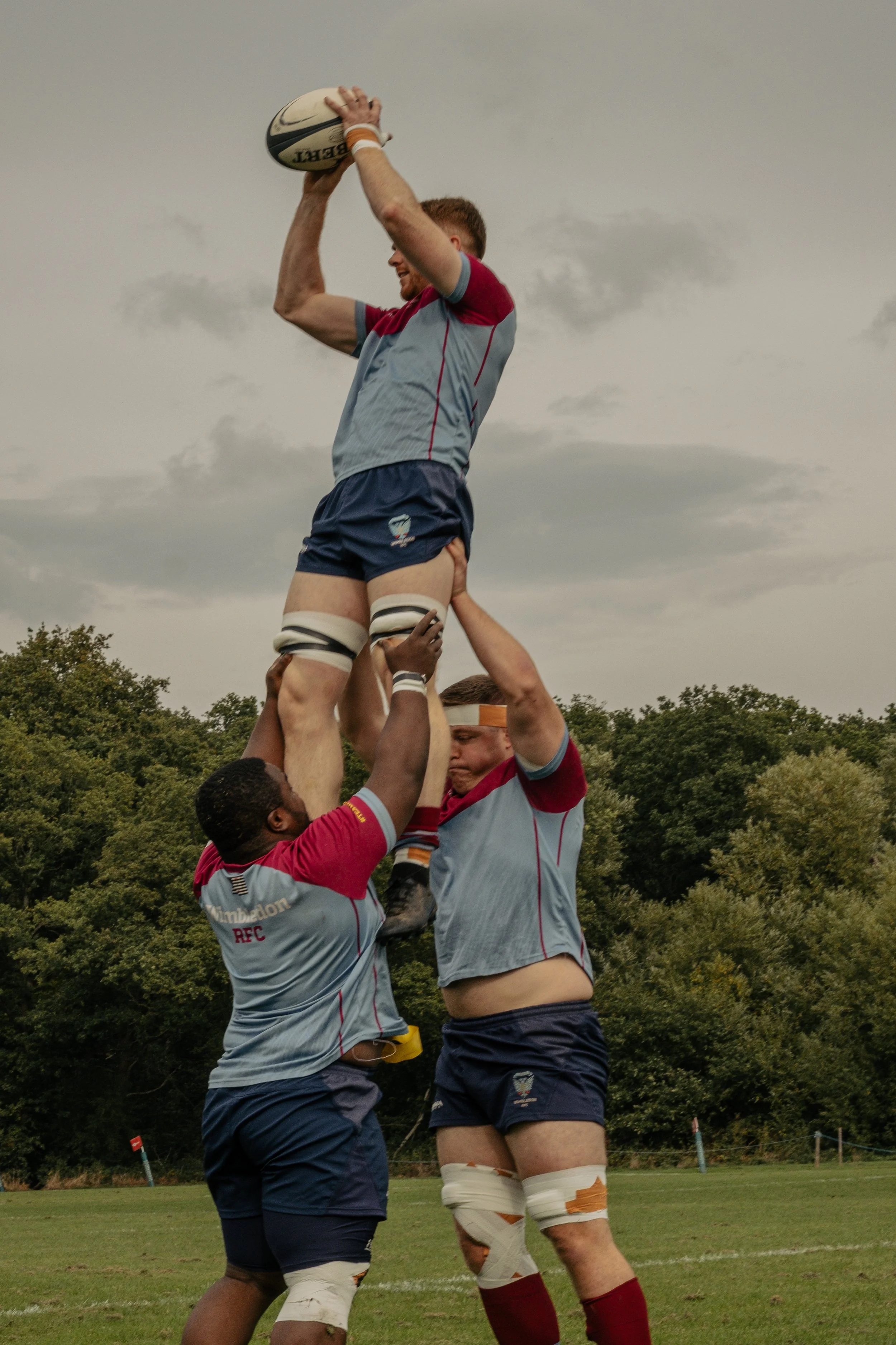 Rugby players lifting a teammate in a line-out during a match, outdoors on a field with trees and cloudy sky in the background.