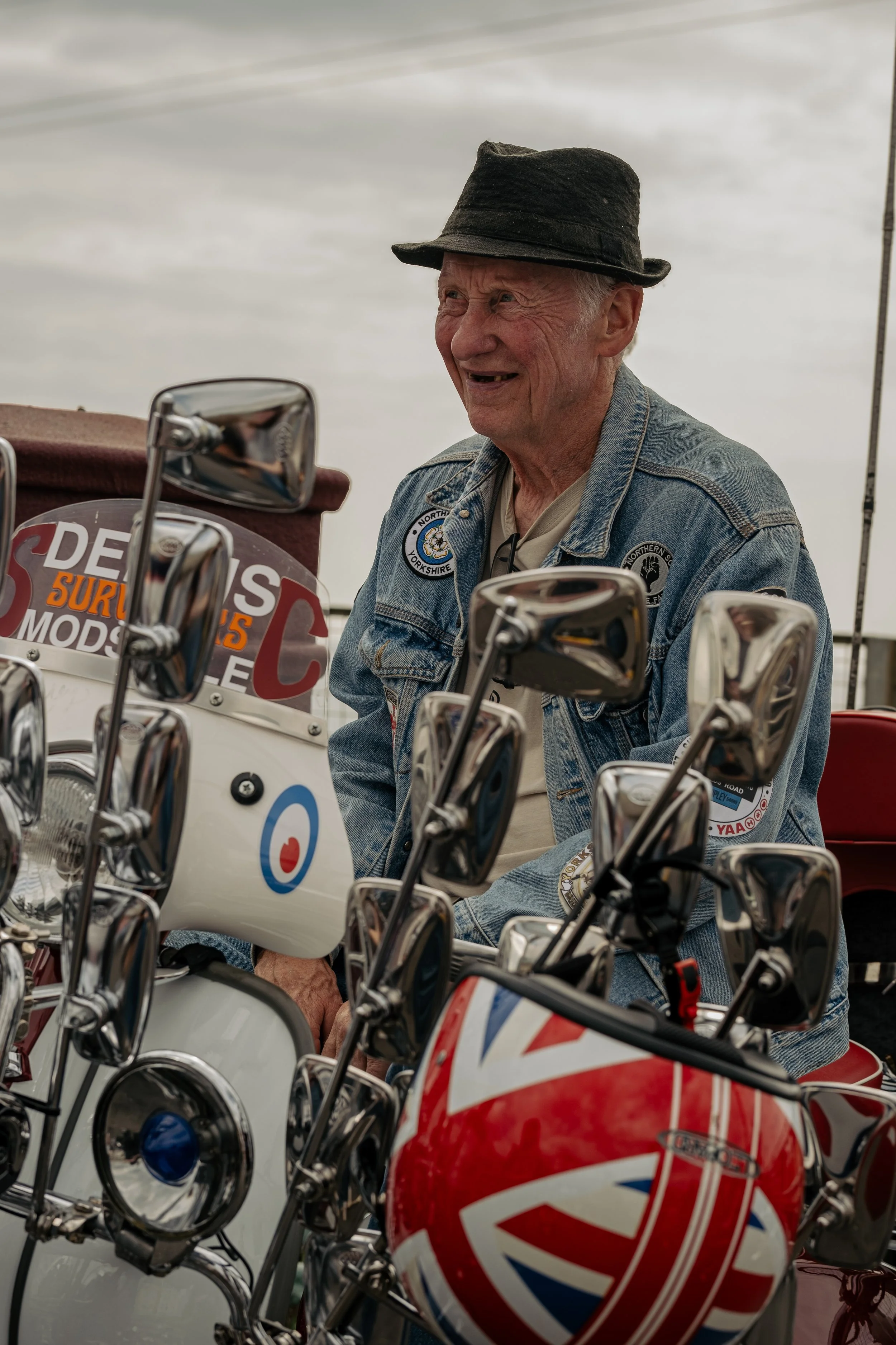 An elderly man with gray hair, wearing a black hat and denim jacket with patches, is smiling and sitting behind a row of vintage motorcycles and scooters decorated with Union Jack and British flag motifs.