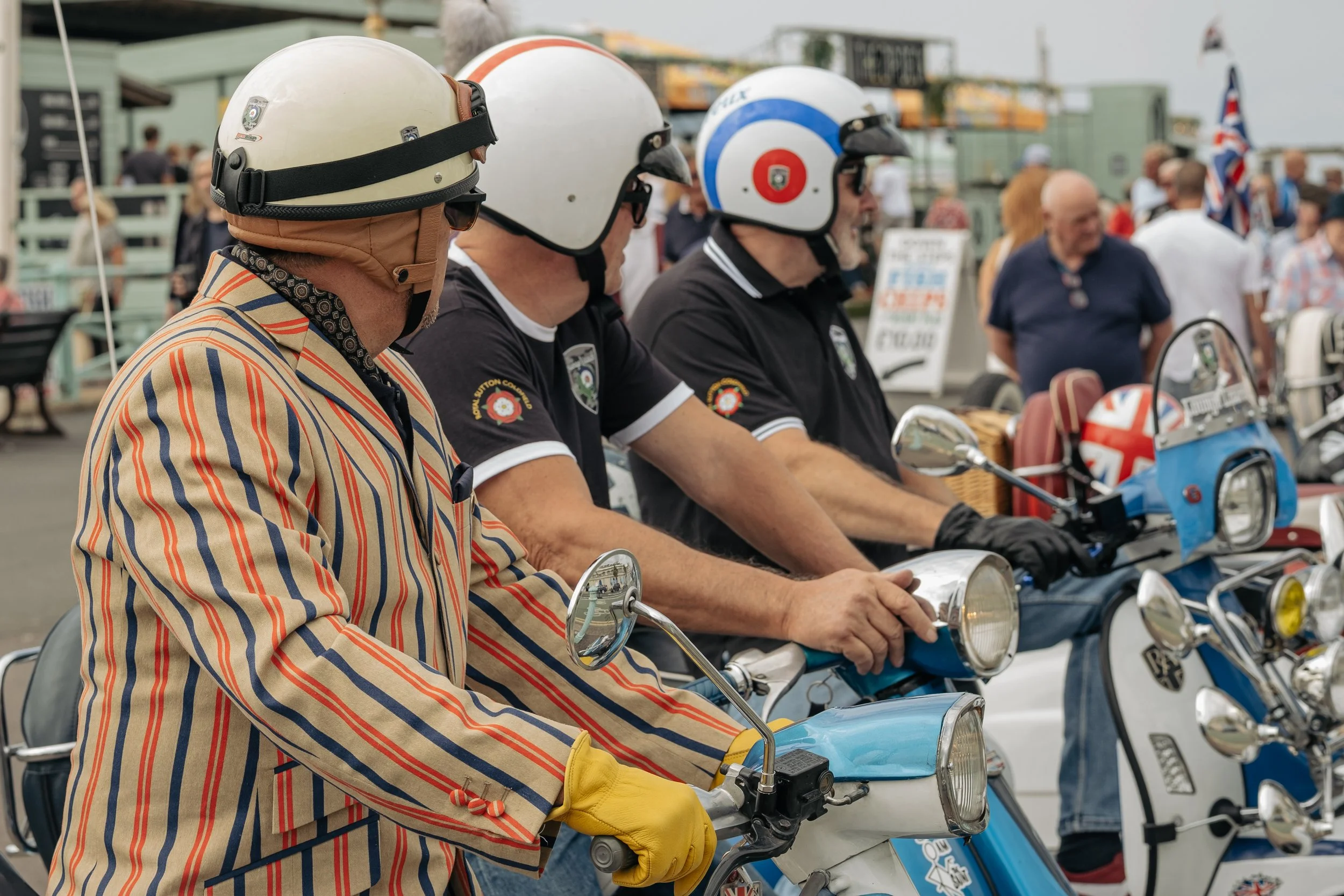 Three men wearing vintage-style helmets and black shirts sit on blue scooters at a gathering, with people in the background, some holding Union Jack flags.