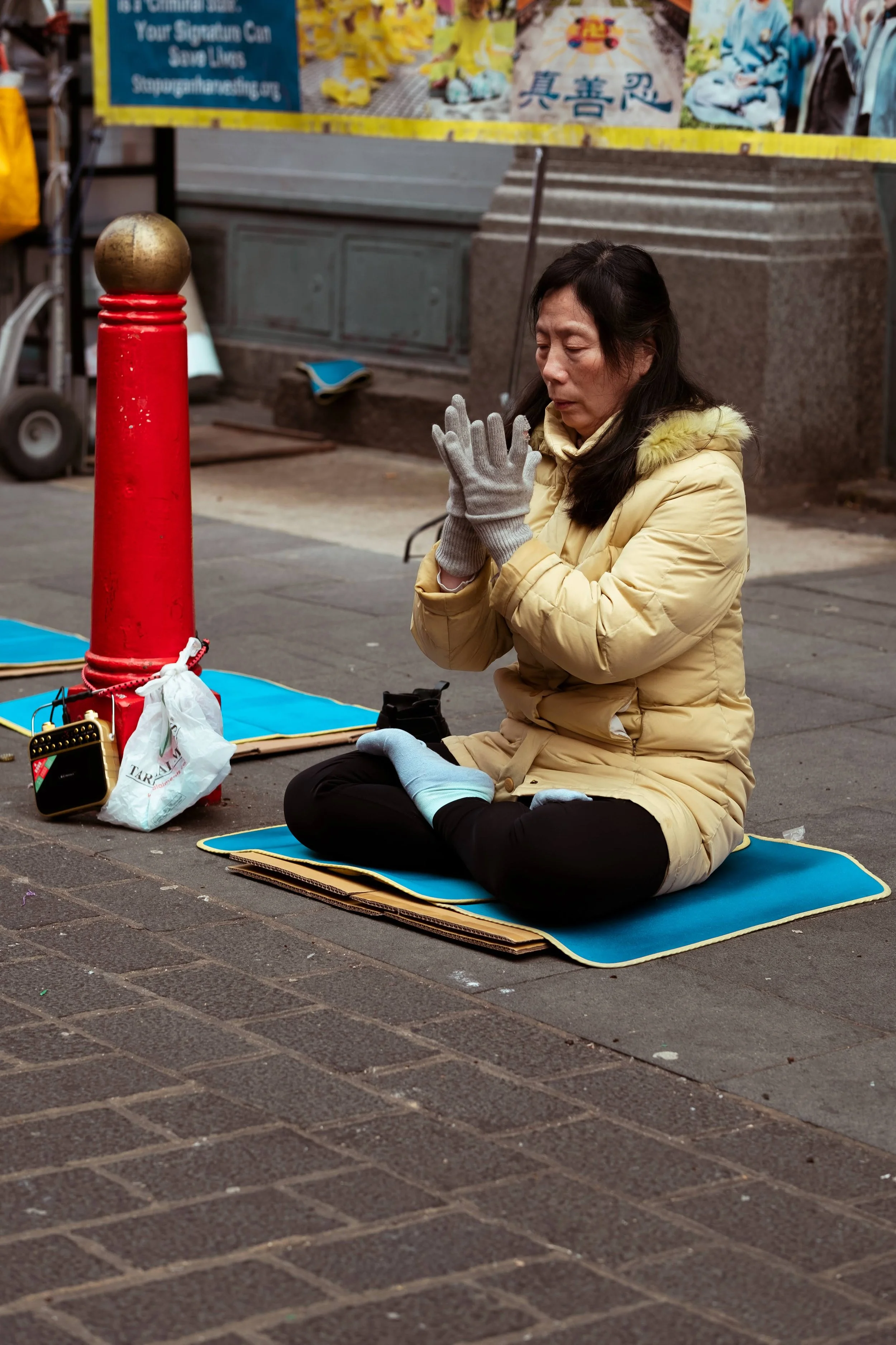 A woman in a yellow coat and gray gloves is kneeling on a blue mat on the sidewalk, praying with her eyes closed and hands clasped. She is surrounded by signs and posters in an urban setting.
