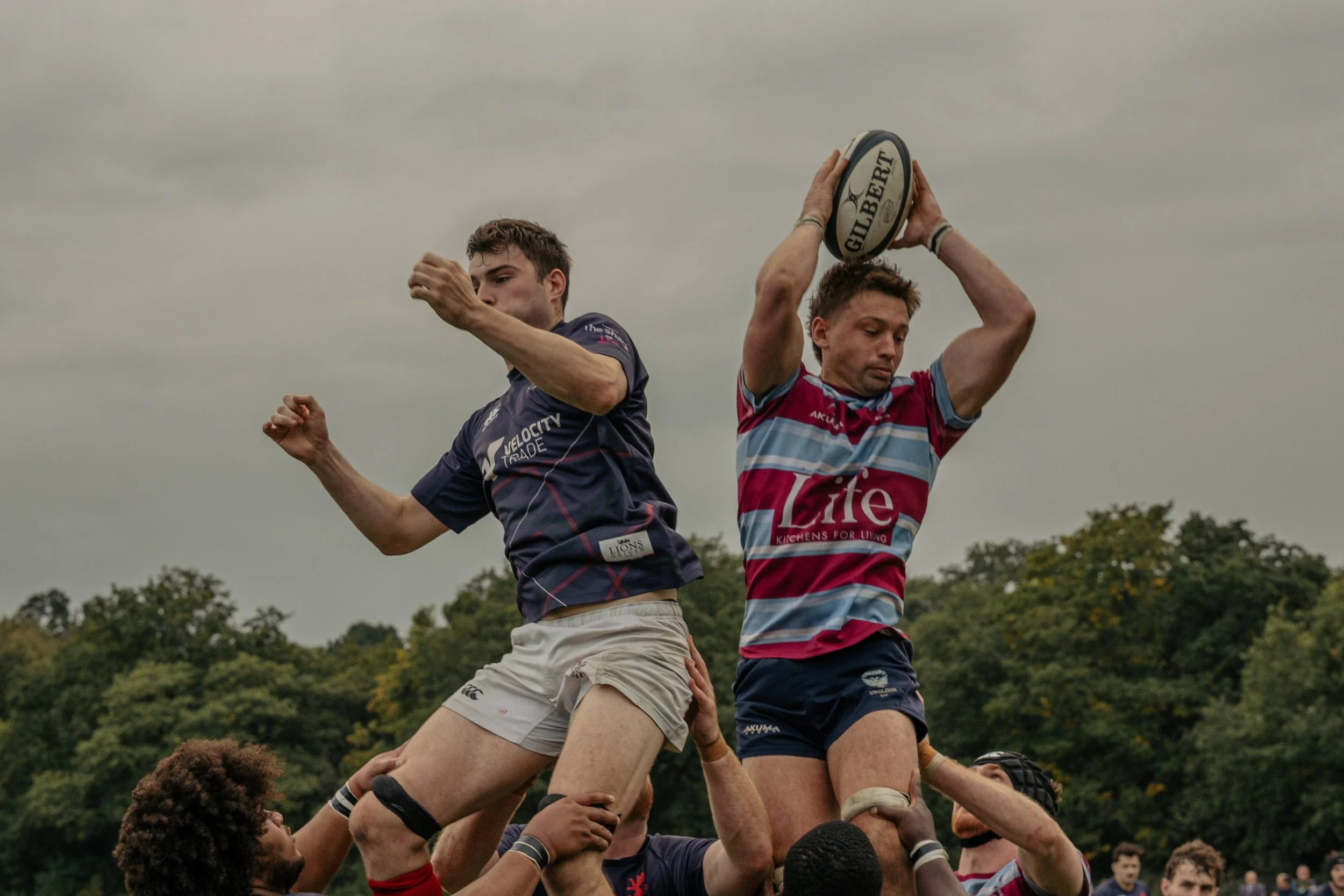 Rugby players competing in a lineout, with one player holding a rugby ball labeled 'Gilbert' while others lift him, against a background of cloudy sky and trees.