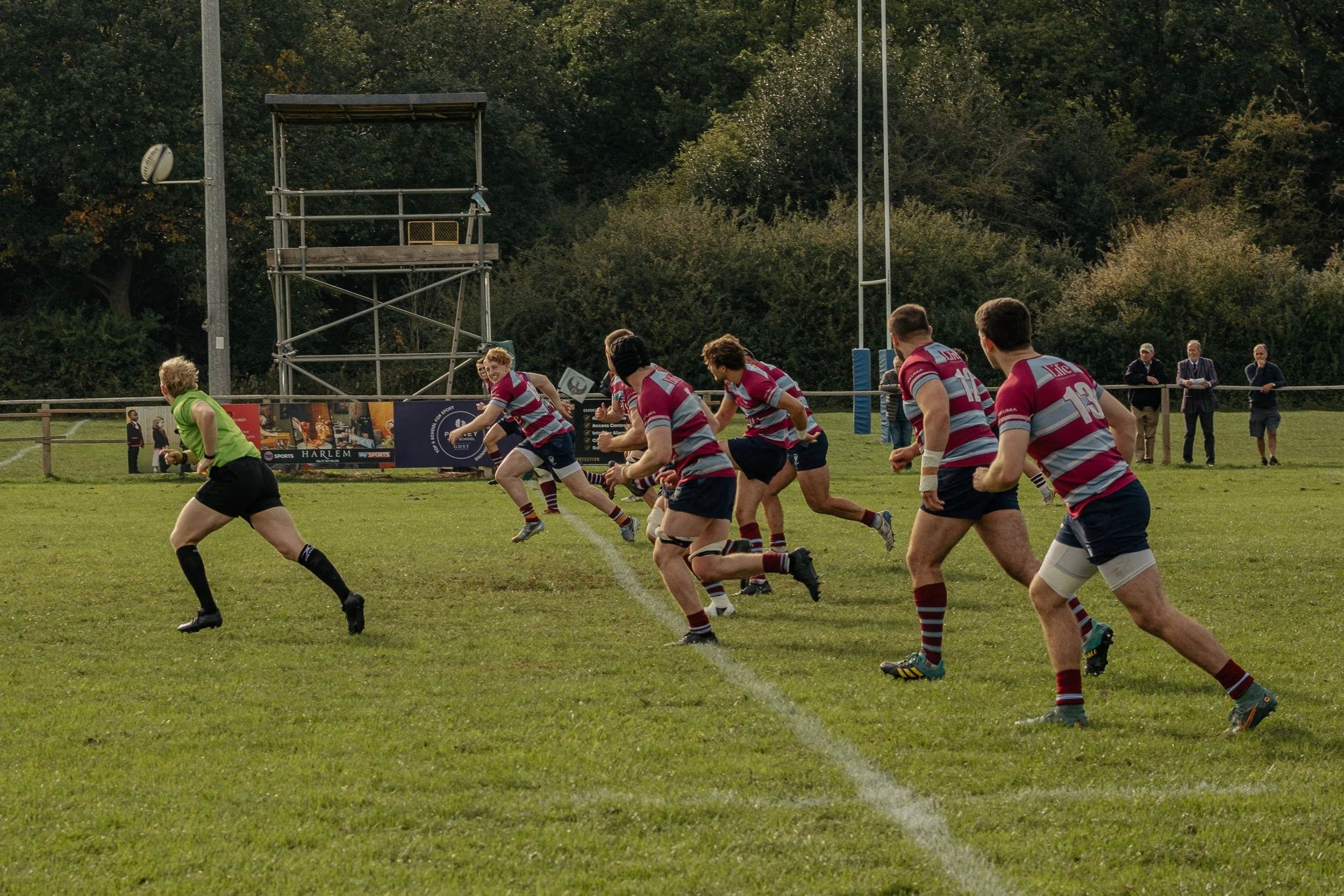 A rugby team in pink and blue uniforms running onto the field with a female referee in a green jersey leading them, in a grassy outdoor stadium surrounded by trees and spectators.
