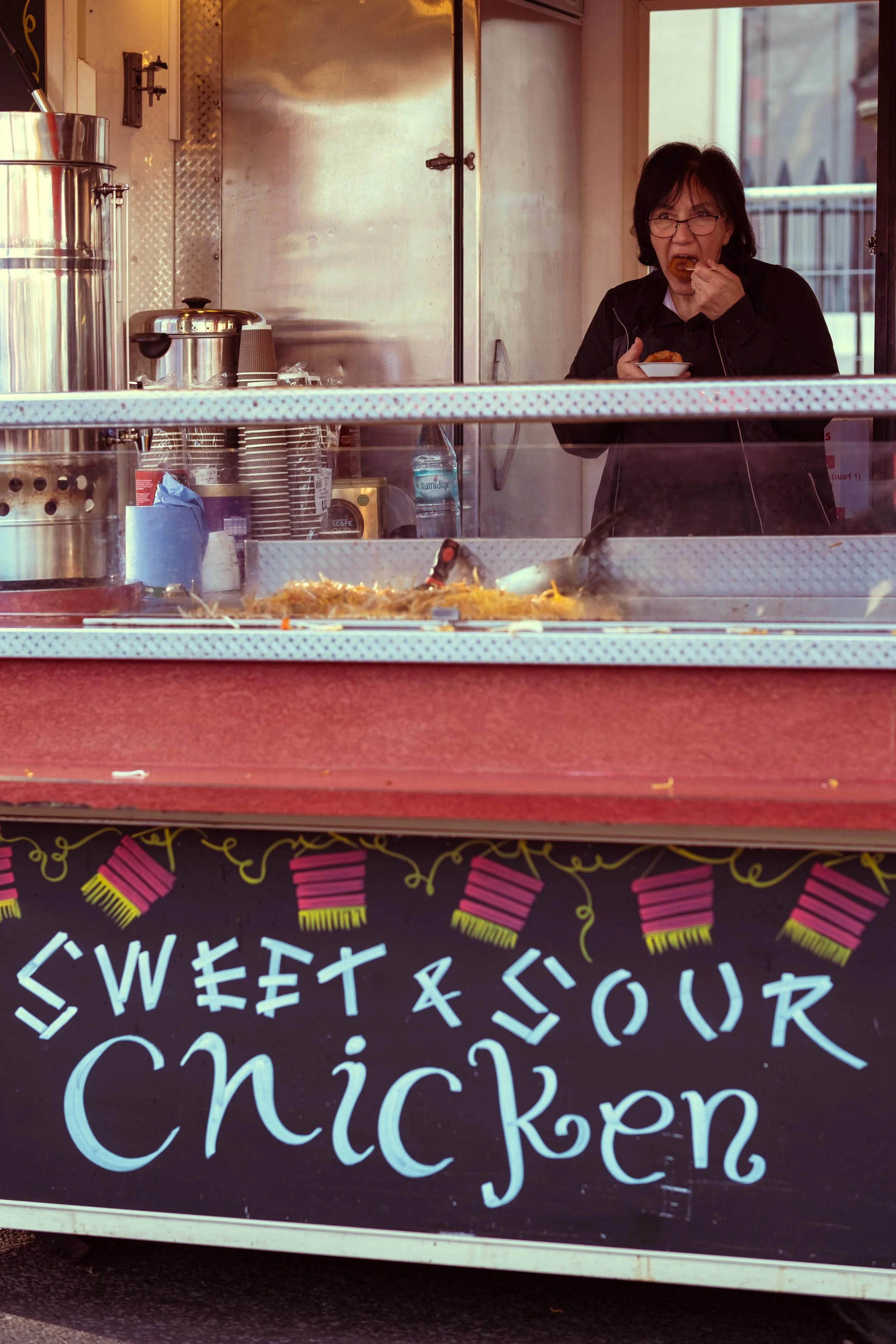A woman eating food inside a chicken stand with a sign that reads 'Sweet & Sour Chicken' and pink and yellow decorations.