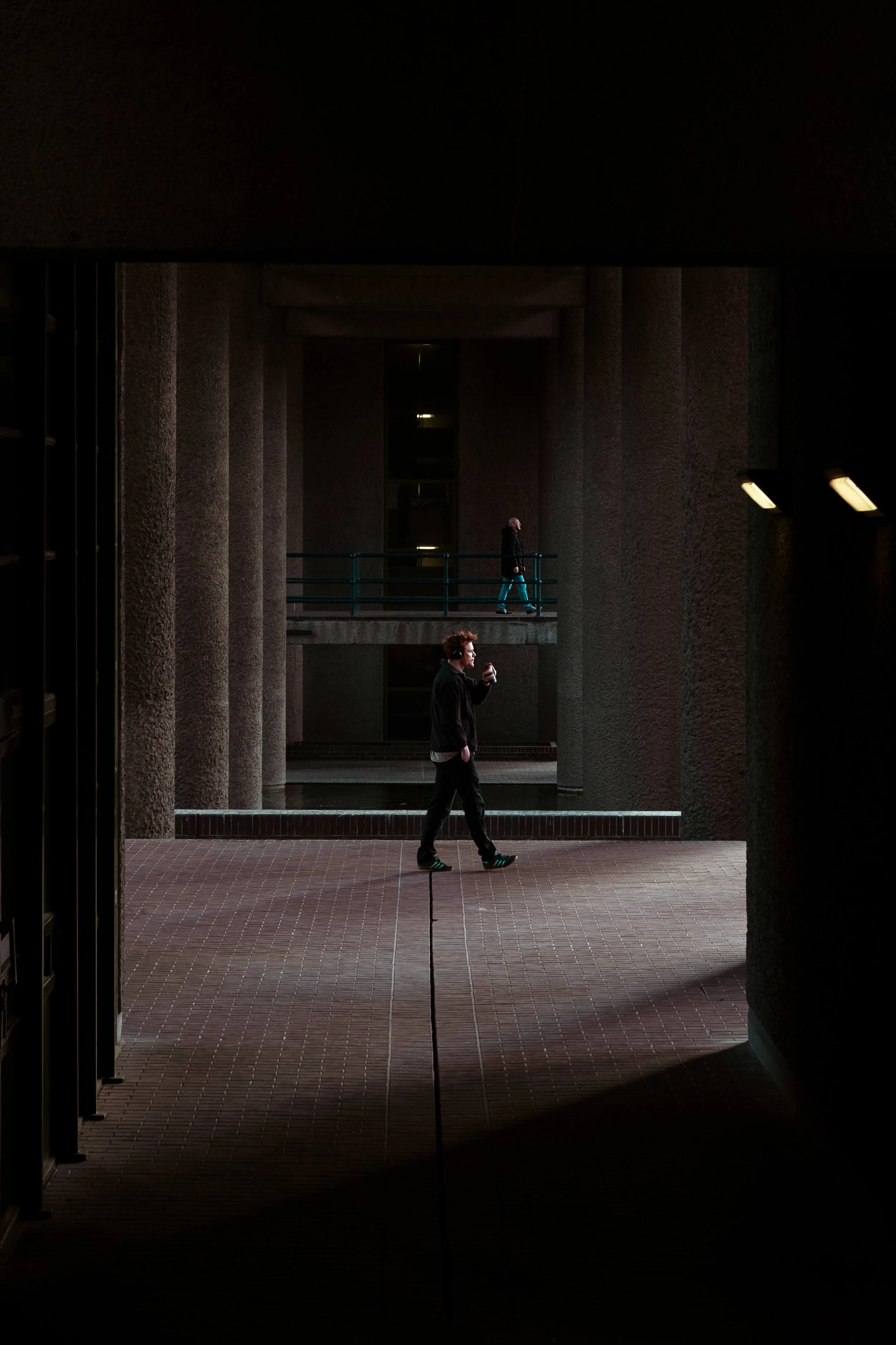 Man walking while talking on cellphone in an urban subway station, with a woman walking on a bridge above.
