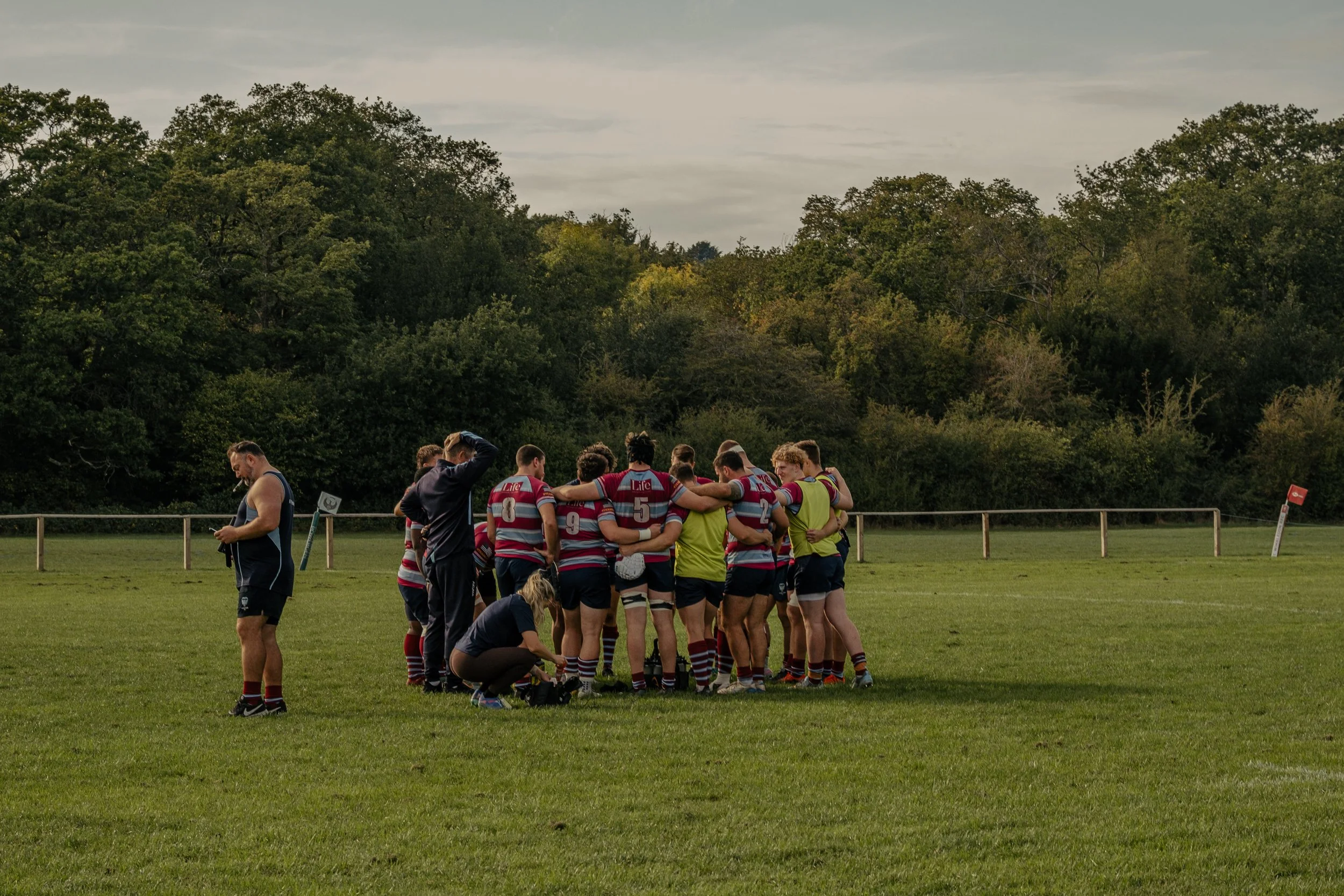 Rugby team in a huddle on a grassy field with trees in the background.