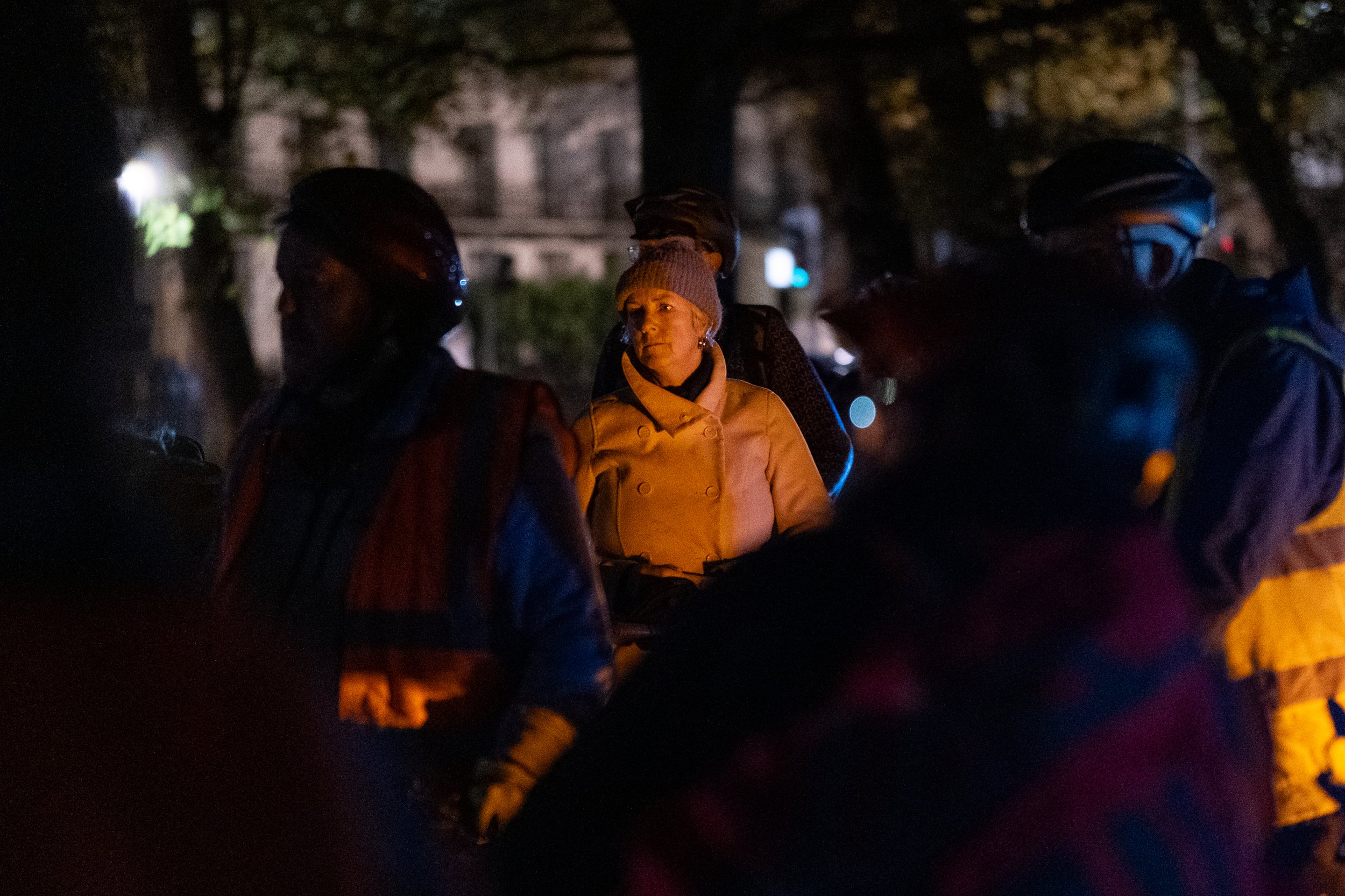 A woman wearing a tan coat and gray knit hat standing among a group of people at night, with some individuals wearing helmets and reflective jackets, in a dark outdoor setting with faint city lights in the background.