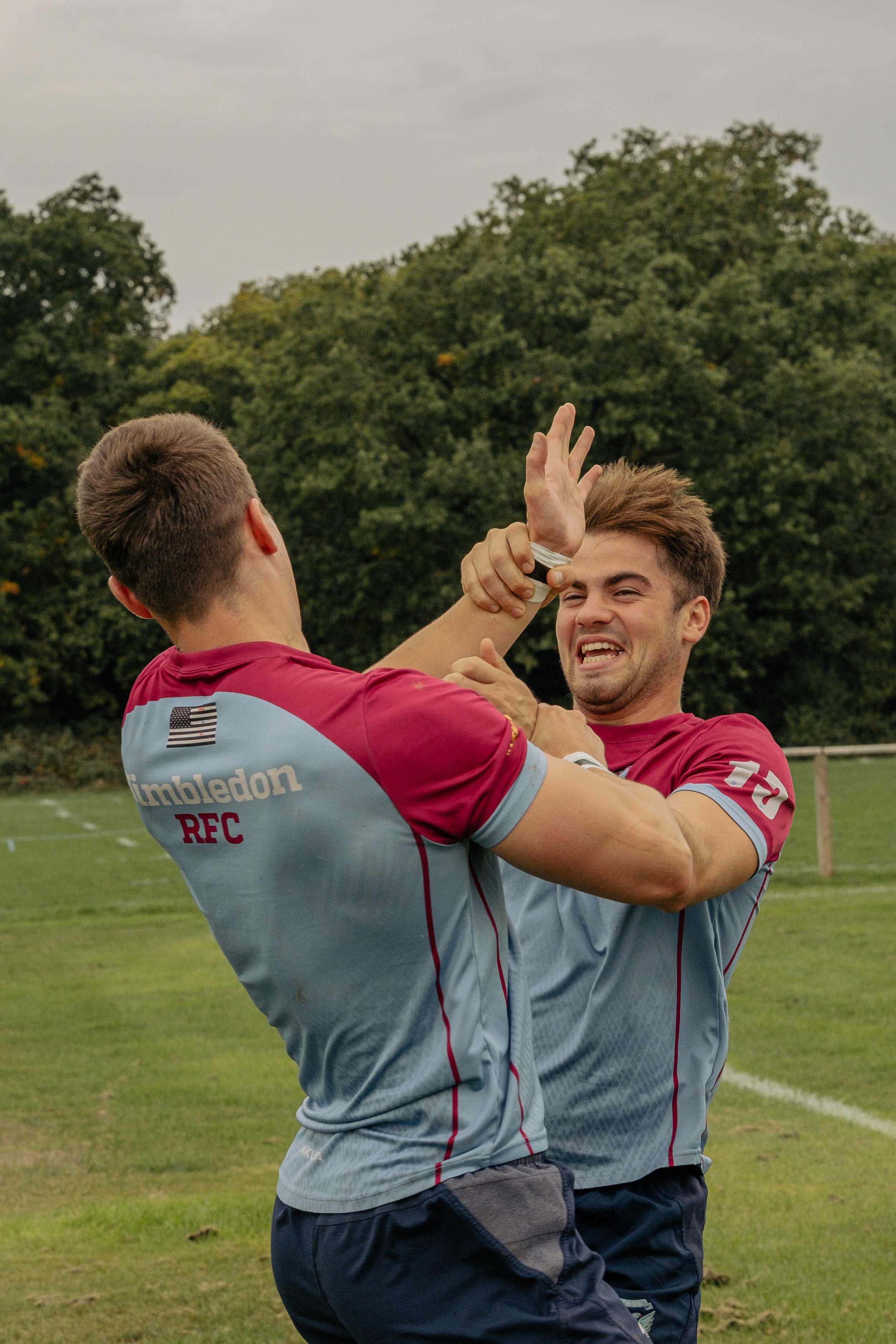 Two rugby players in maroon and light blue jerseys are engaged in a physical confrontation, with one player grabbing the other's face and neck as they struggle on a grassy field.