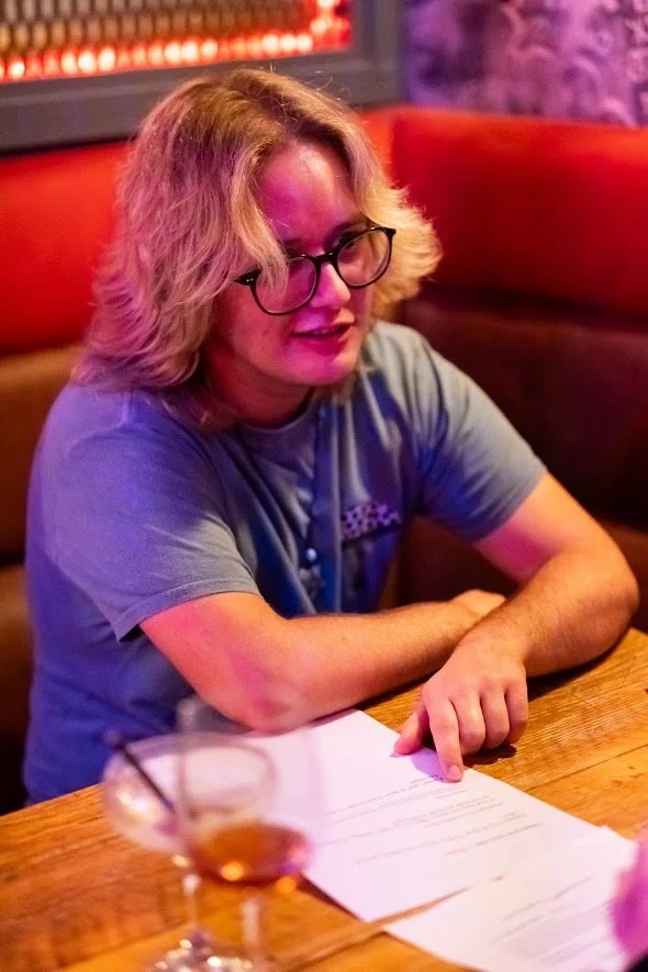 A woman with blonde curly hair and glasses sitting at a wooden table in a dimly lit restaurant or bar, pointing at a menu or paper, with a drink in a glass and a straw in the foreground.