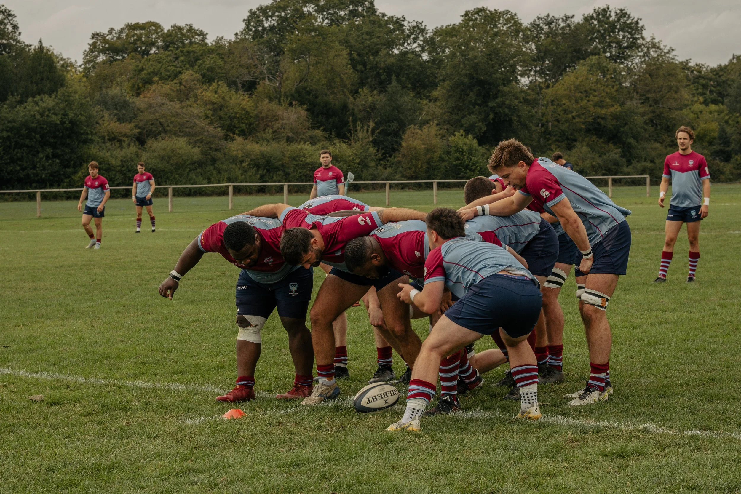 A rugby team in red and blue uniforms huddles on a grassy field, preparing for a play, with other players standing in the background, surrounded by trees.