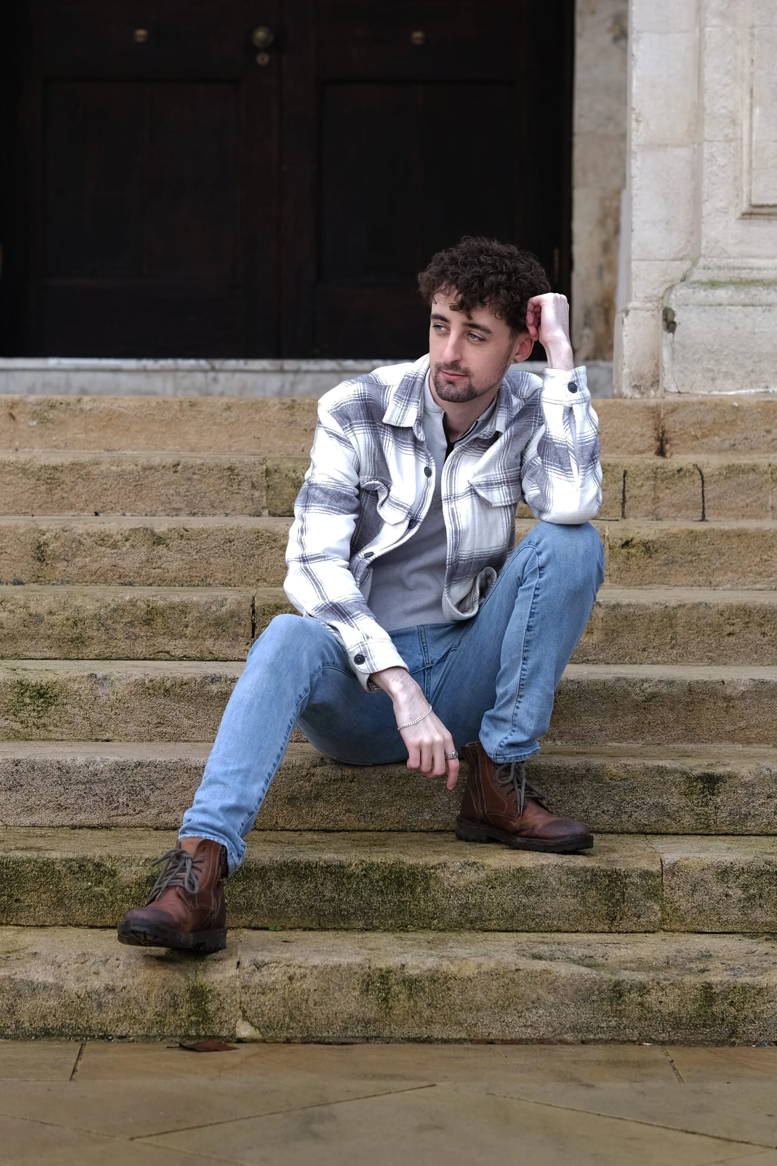 A young man with curly hair and a beard sitting on stone steps in front of a large dark wooden door, wearing a plaid shirt, gray t-shirt, blue jeans, and brown boots, looking thoughtfully to the side.