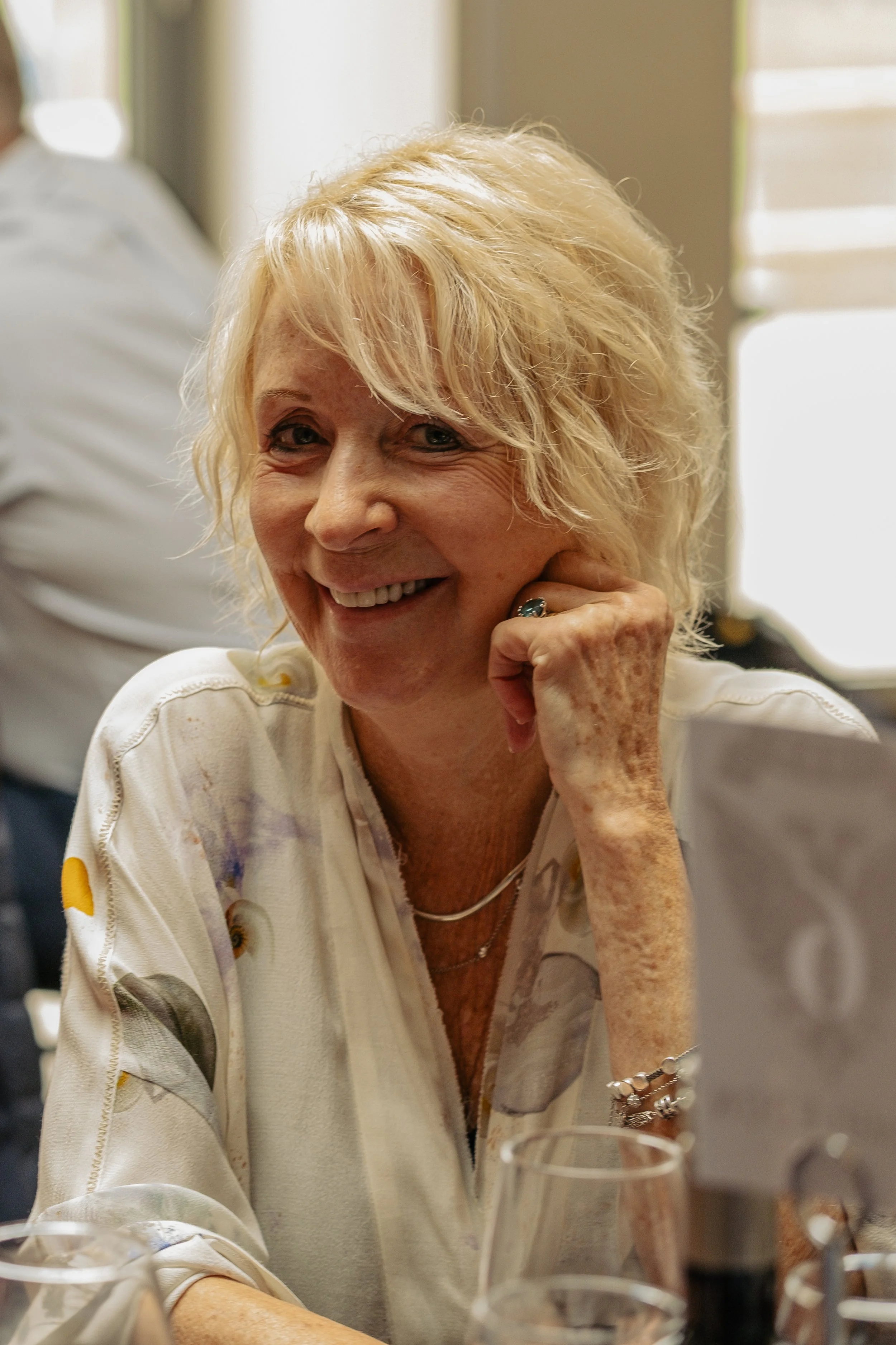 A smiling woman with blonde hair sitting at a table, resting her chin on her hand, wearing a white shirt with a pattern.
