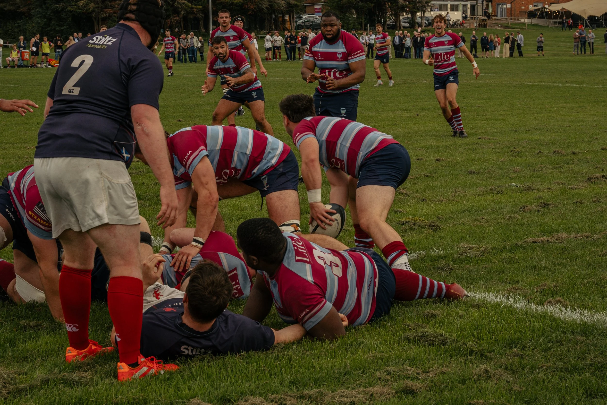 Rugby players in a scrum on a grassy field, with spectators watching in the background.
