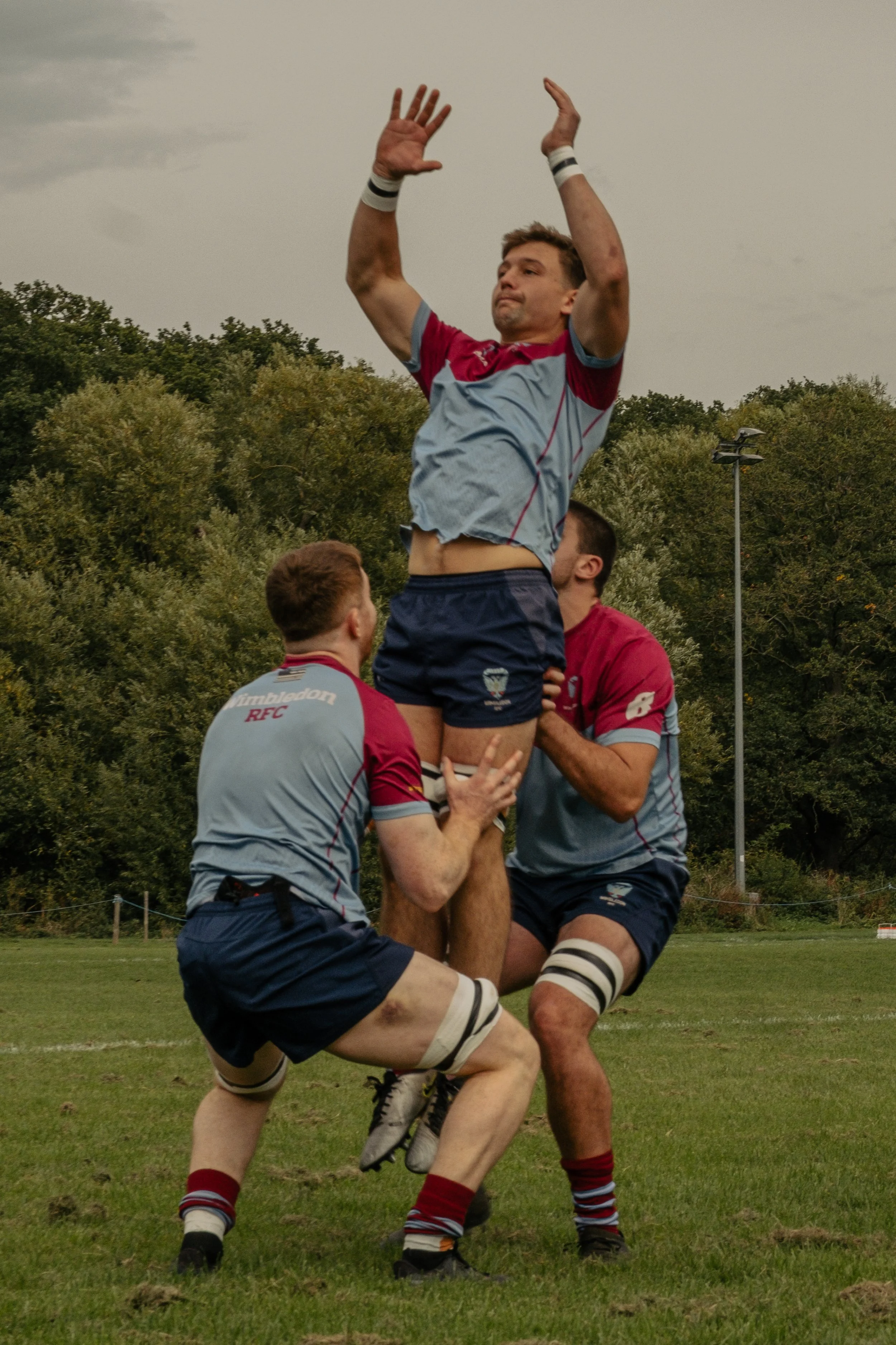 Three rugby players in uniform lifting a teammate for a lineout during a game on a grassy field.