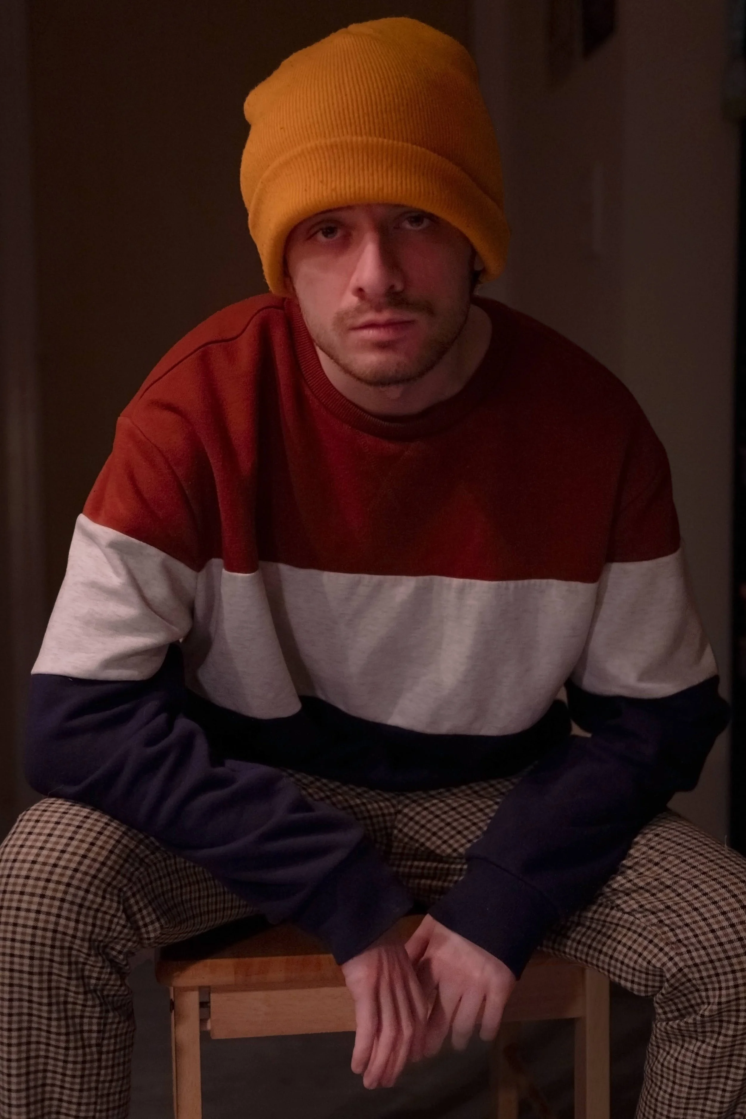A young man wearing a bright yellow beanie, a color-blocked sweatshirt with red, white, and navy sections, and checkered pants, sitting on a wooden chair indoors with dim lighting.