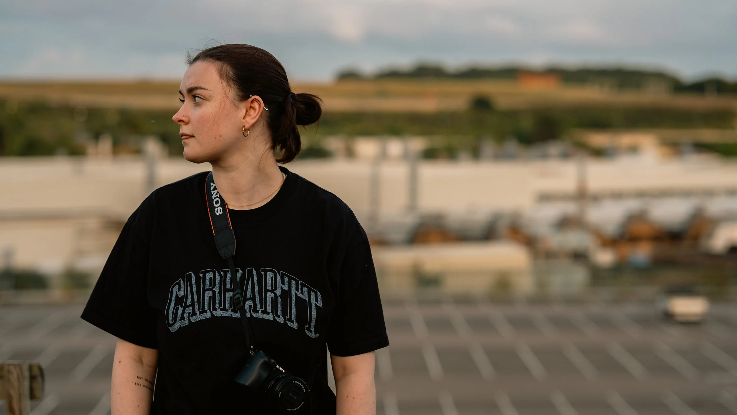 A young woman with brown hair tied back, wearing a black Carhartt t-shirt and a Sony camera around her neck, standing outdoors with a blurred background of a parking lot, buildings, and green hills.